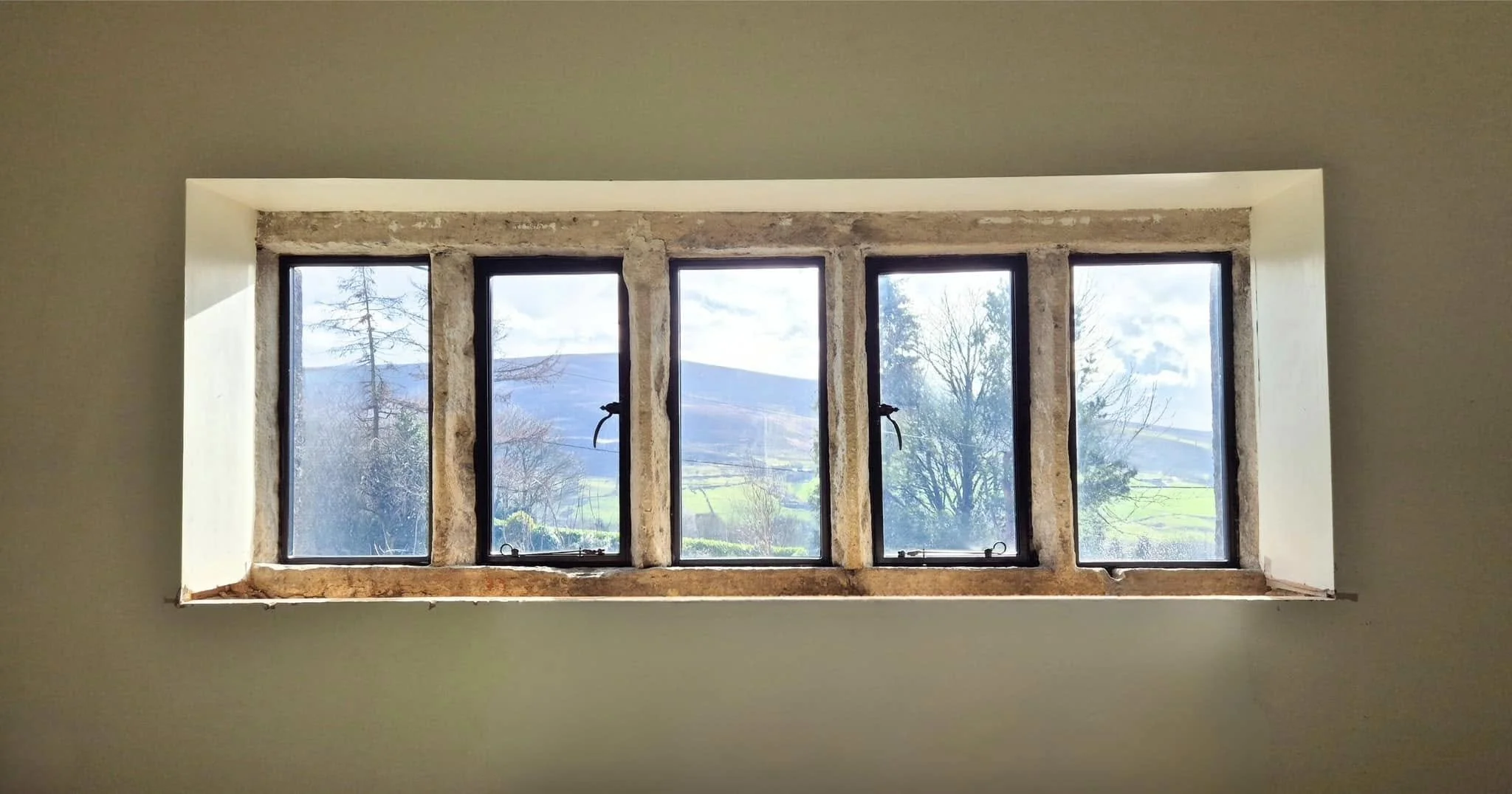 A rectangular window with five panes, set in an unfinished wall, revealing a landscape with trees, hills, and a partly cloudy sky outside.