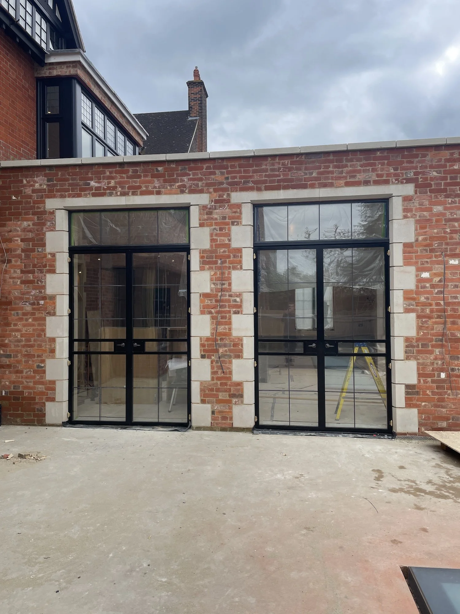 Two large black-framed glass doors installed in a brick building with a concrete floor in front of them, under a partially cloudy sky.