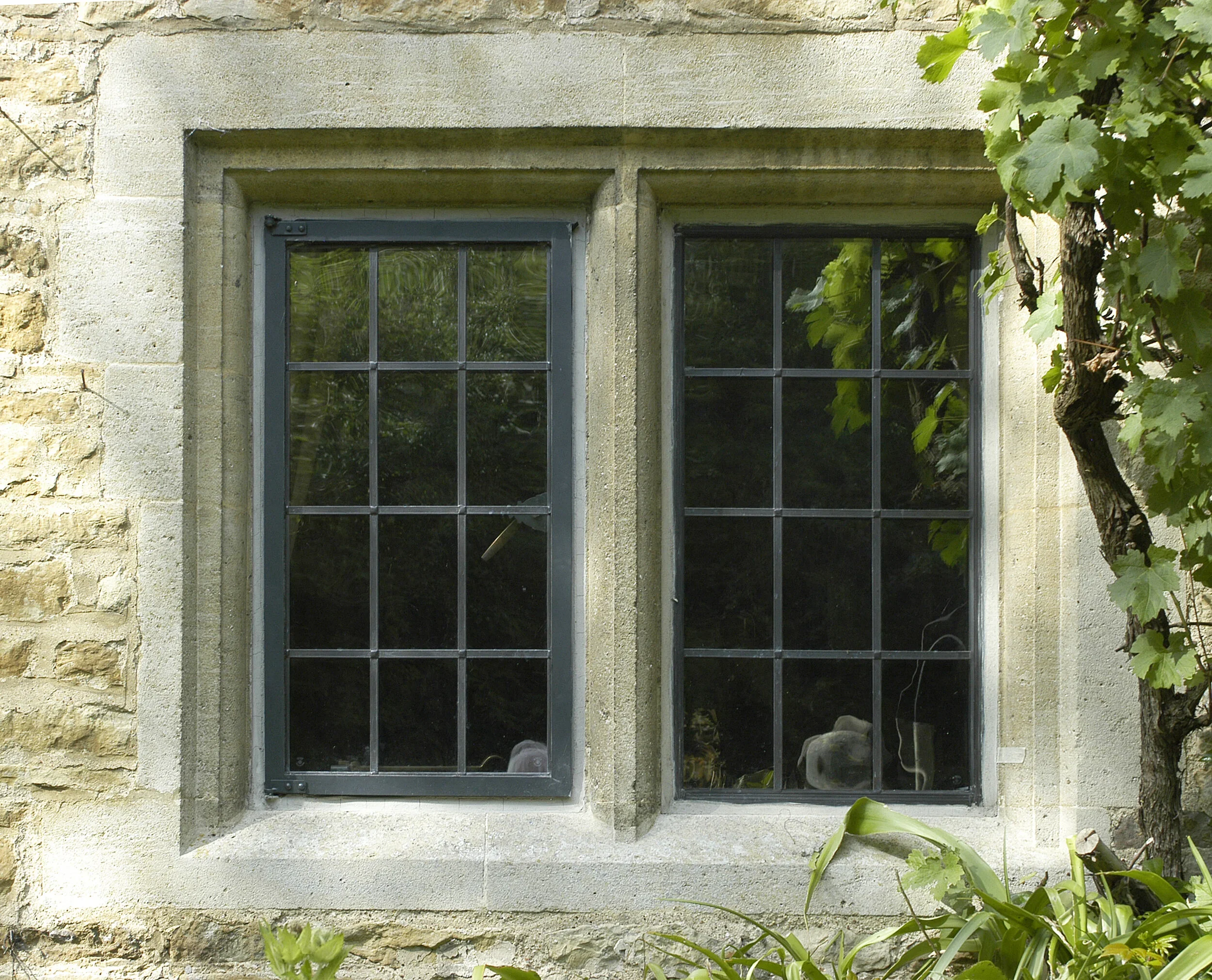 A stone house window with black metal grilles, surrounded by green leaves and plants.