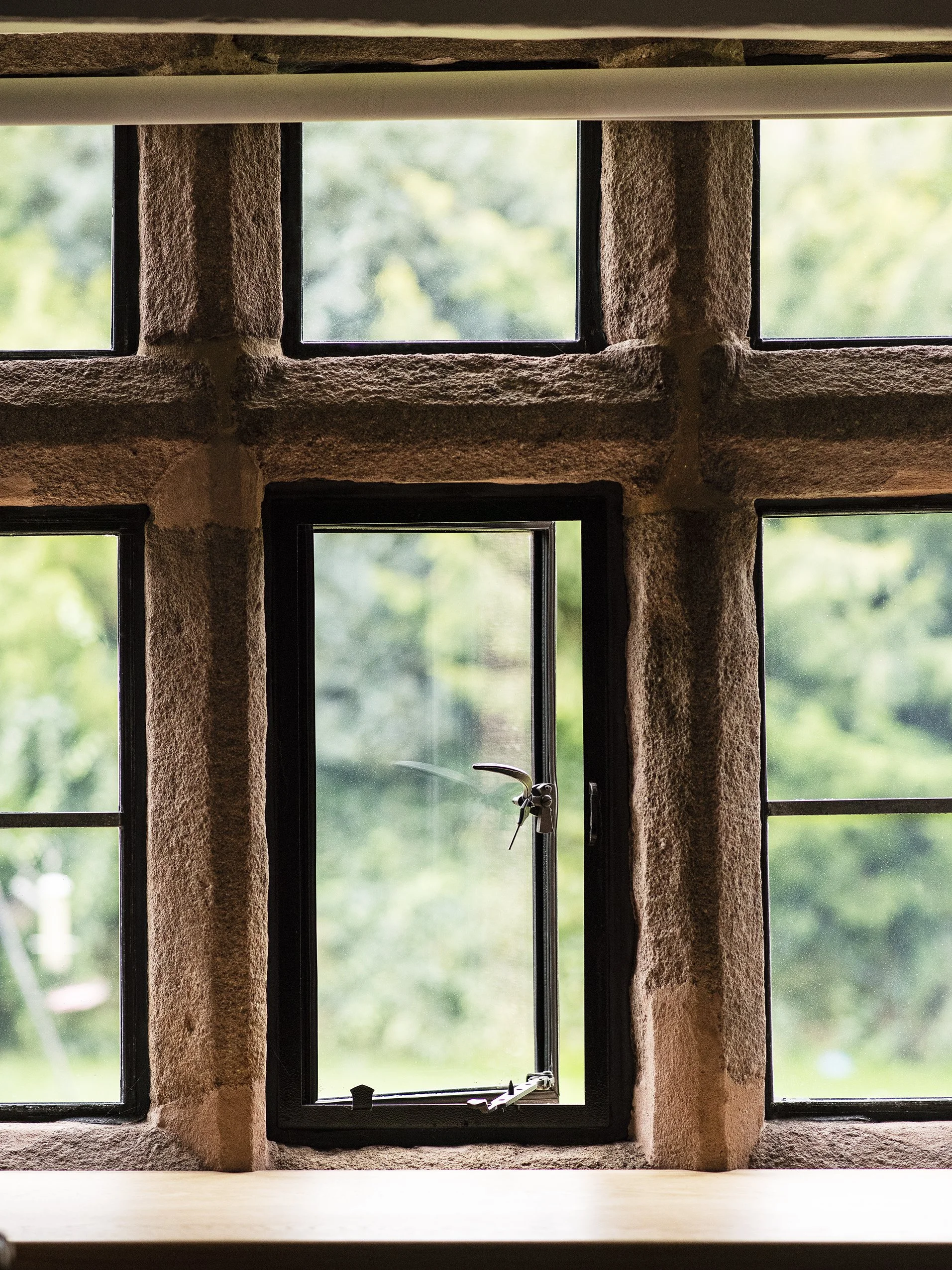 A small window with a black frame, set into a red brick wall, with a latch and a handle turned outward, overlooking a green, leafy outdoor area.