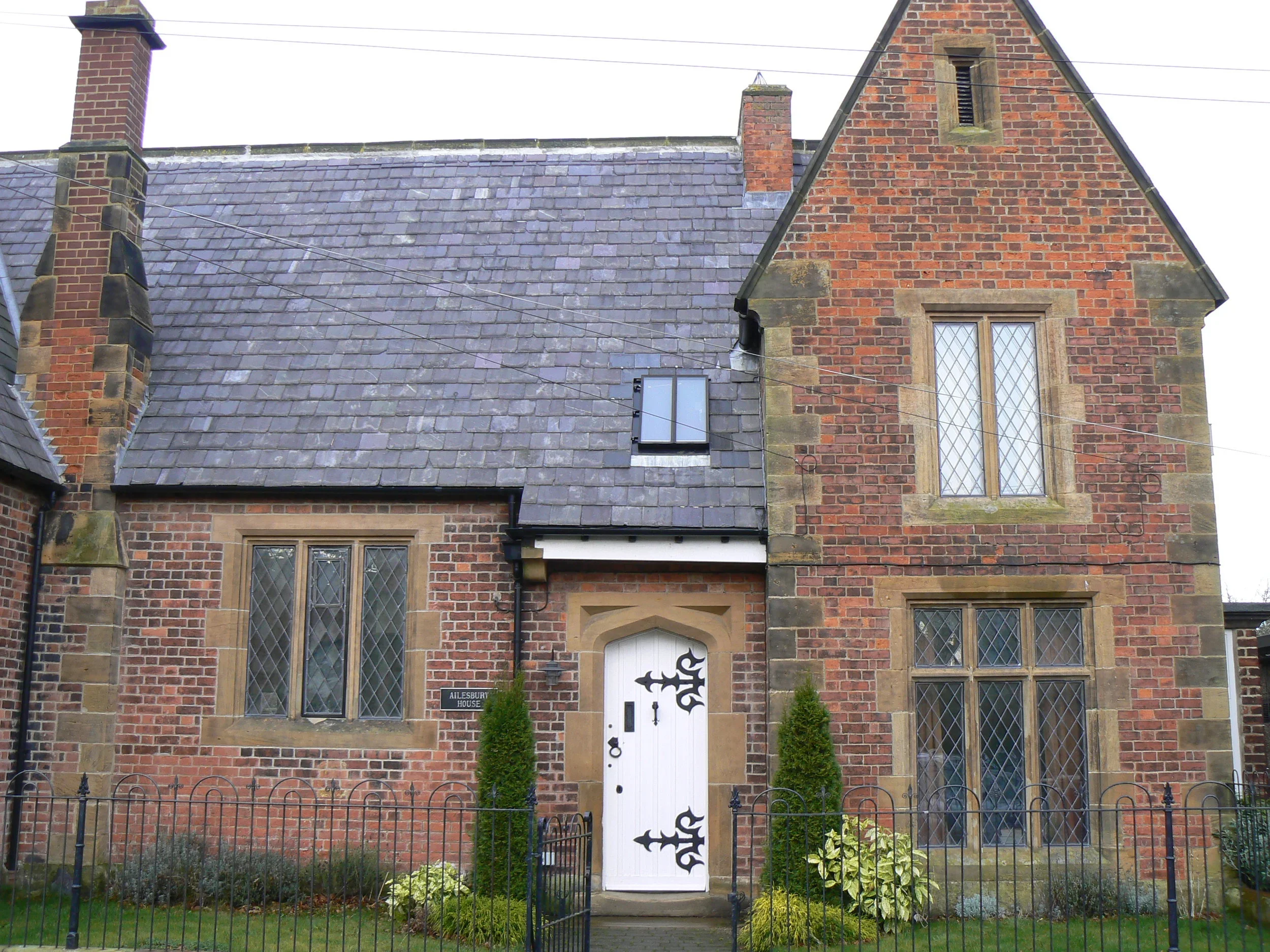 A brick house with a slate roof, a white front door with black decorative hinges, tall narrow windows with lattice glass, two small trees flanking the door, and a low black metal fence in front.