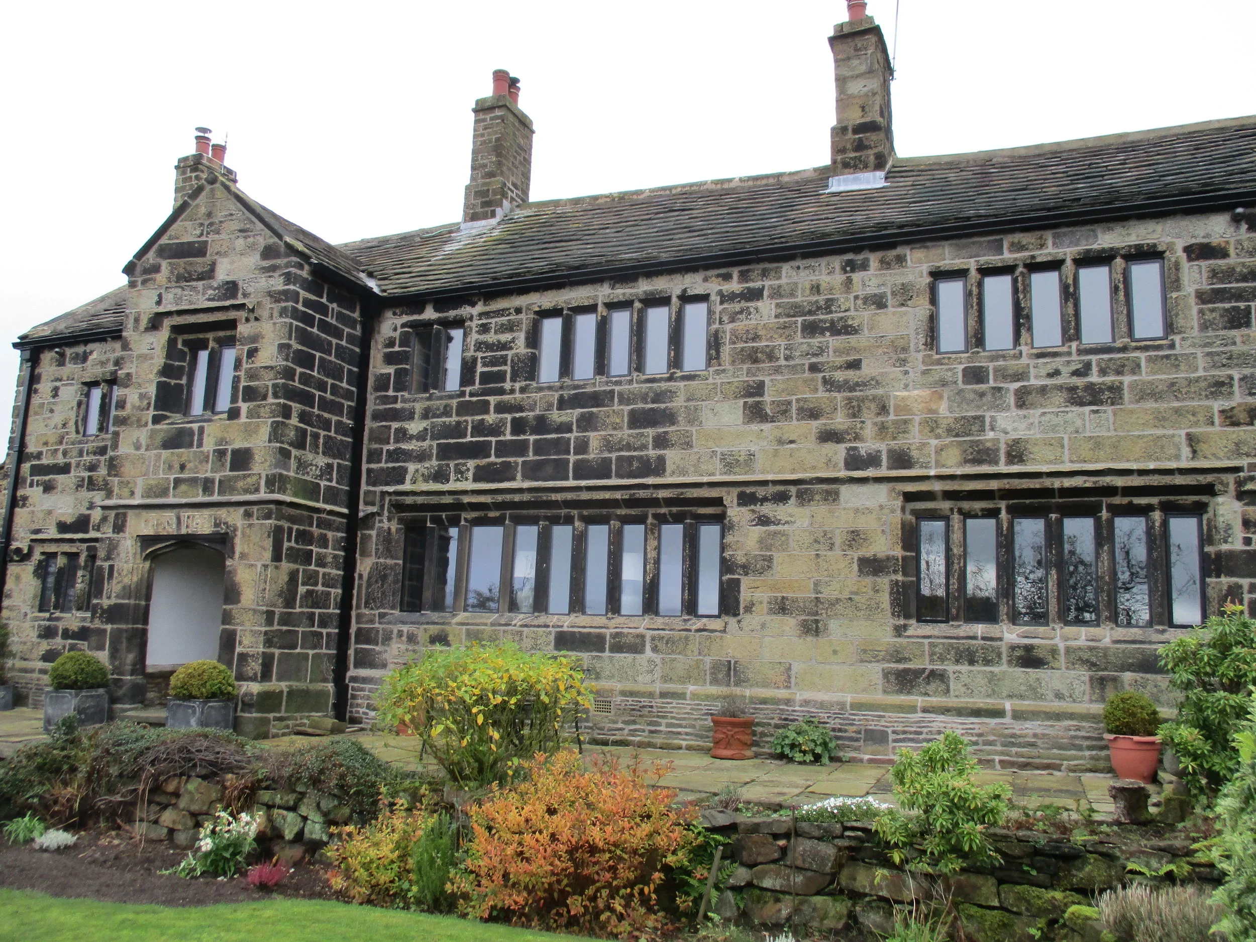 A historic stone house with multiple chimneys, small windows, and a garden in front with various plants and pots.