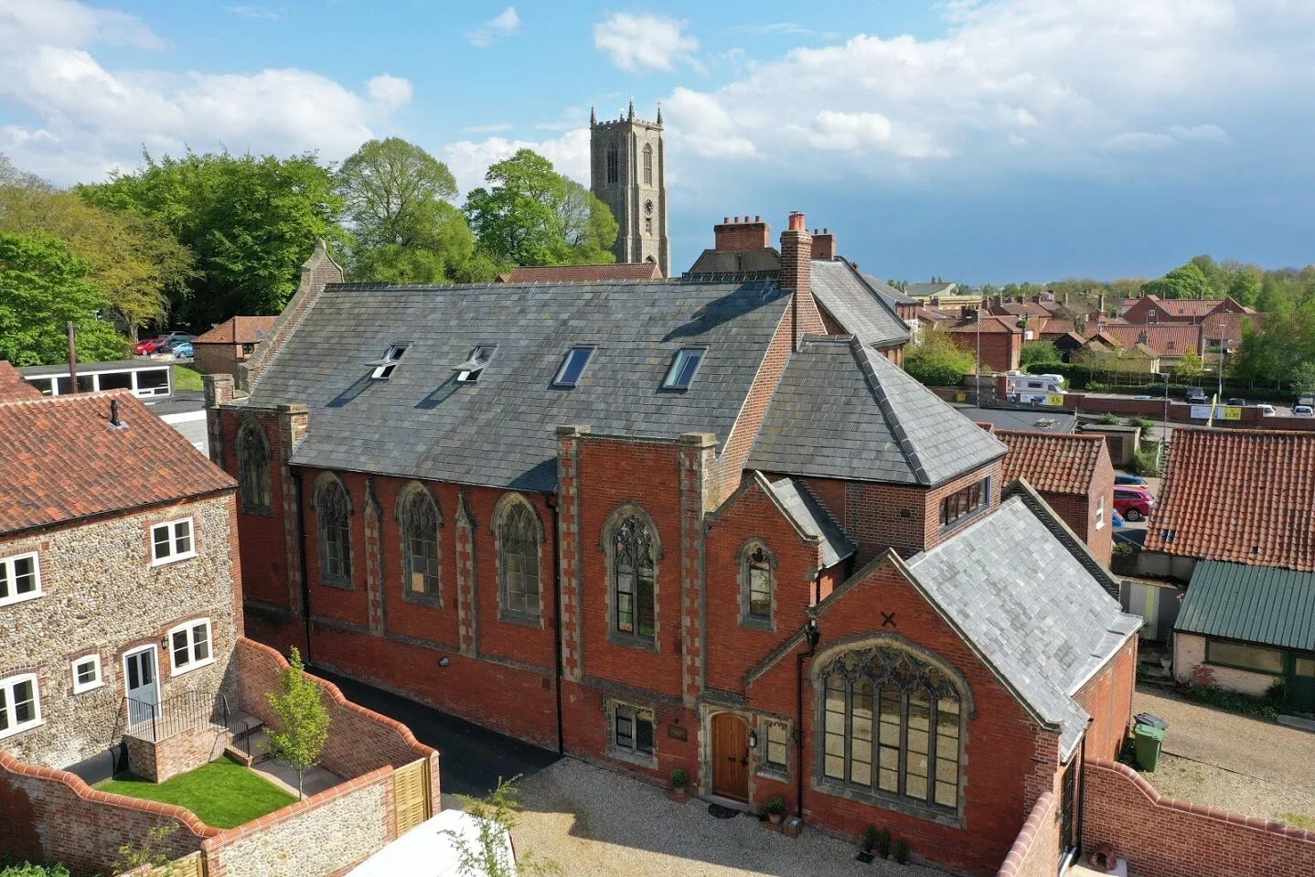 Aerial view of a historic red brick church with arched windows, situated in a small town with greenery and other buildings under a partly cloudy sky.