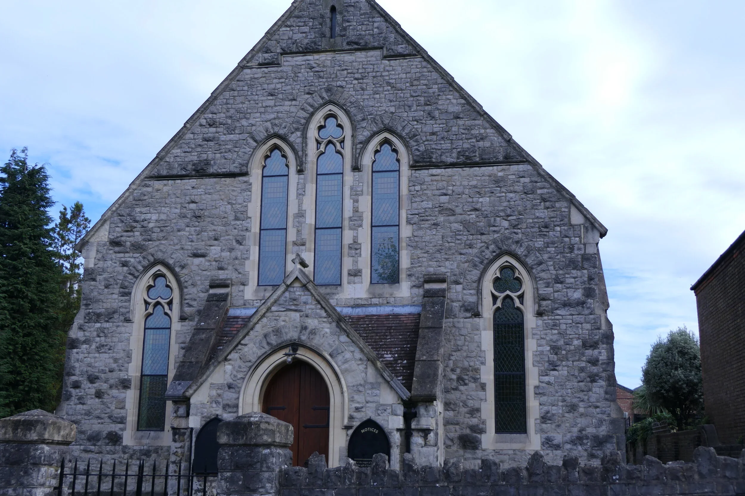 A stone church with Gothic-style pointed arched windows and a wooden door, surrounded by a stone fence and greenery, under a cloudy sky.