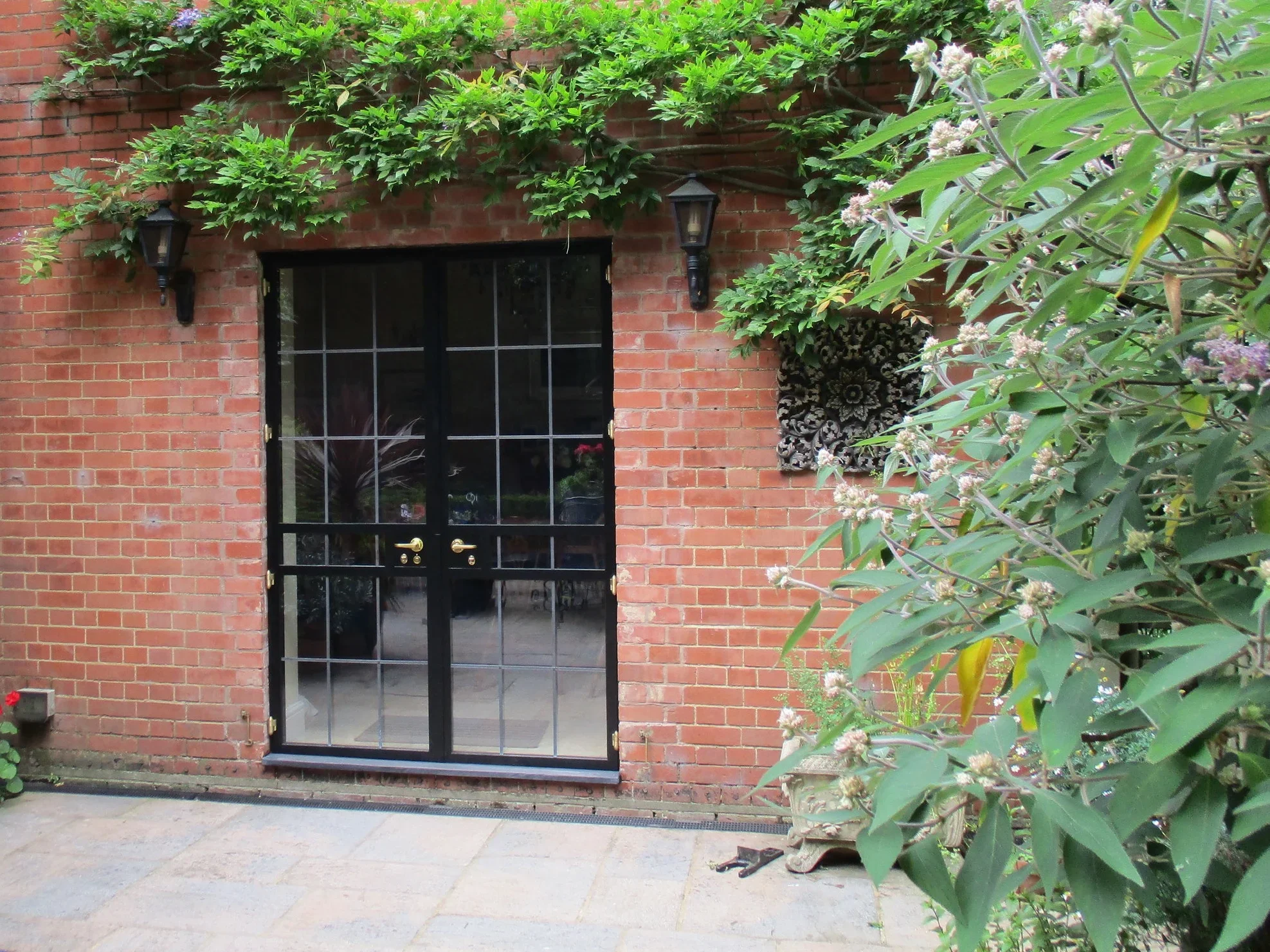 A brick wall with black-framed glass double doors, surrounded by green plants and flowers, with wall-mounted lanterns on either side of the doors.