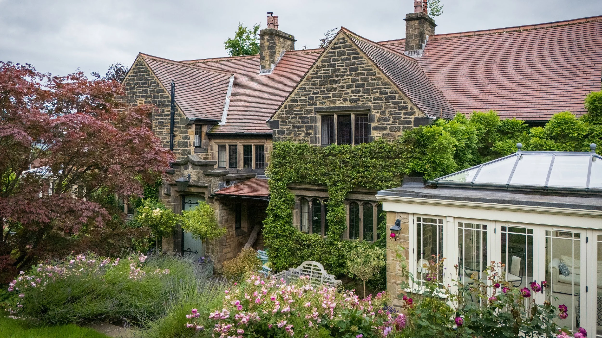 A large, historic stone house with a red-tiled roof surrounded by a lush, colorful garden and a conservatory with glass panels.
