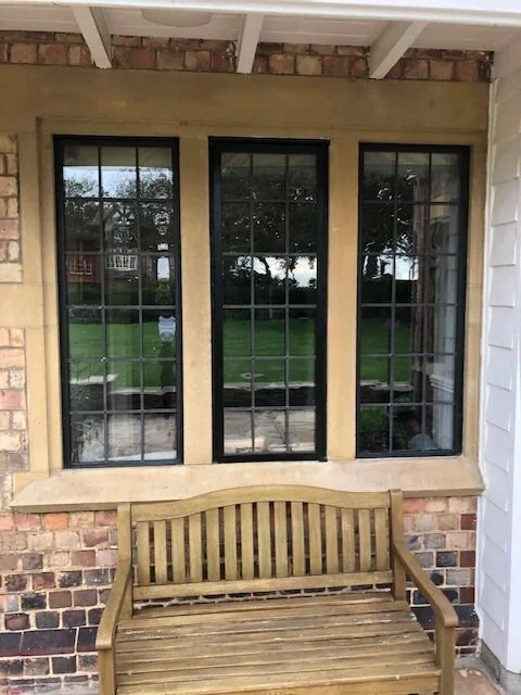 Three tall black-framed windows with multiple panes set into a brick wall under a white porch ceiling, with a wooden bench beneath them and a grassy yard with trees visible outside.