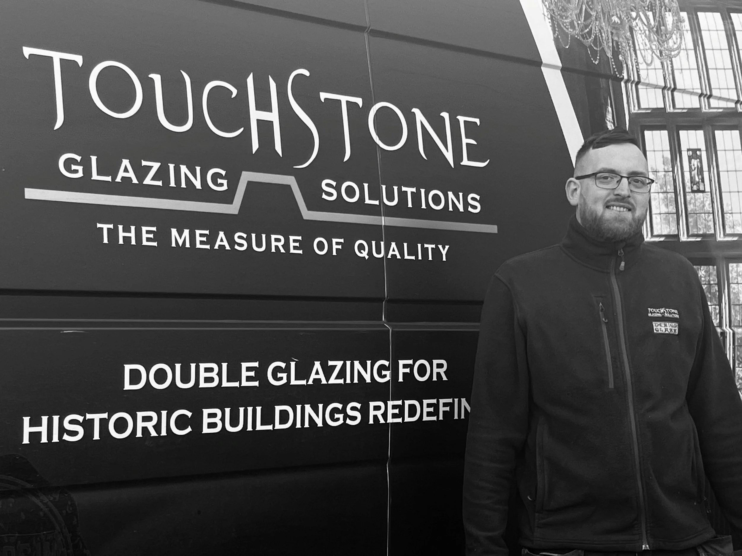 A man with glasses and a beard standing in front of a sign for Touchstone Glazing Solutions, a window or glass company, inside a building with large windows and ornate chandeliers.