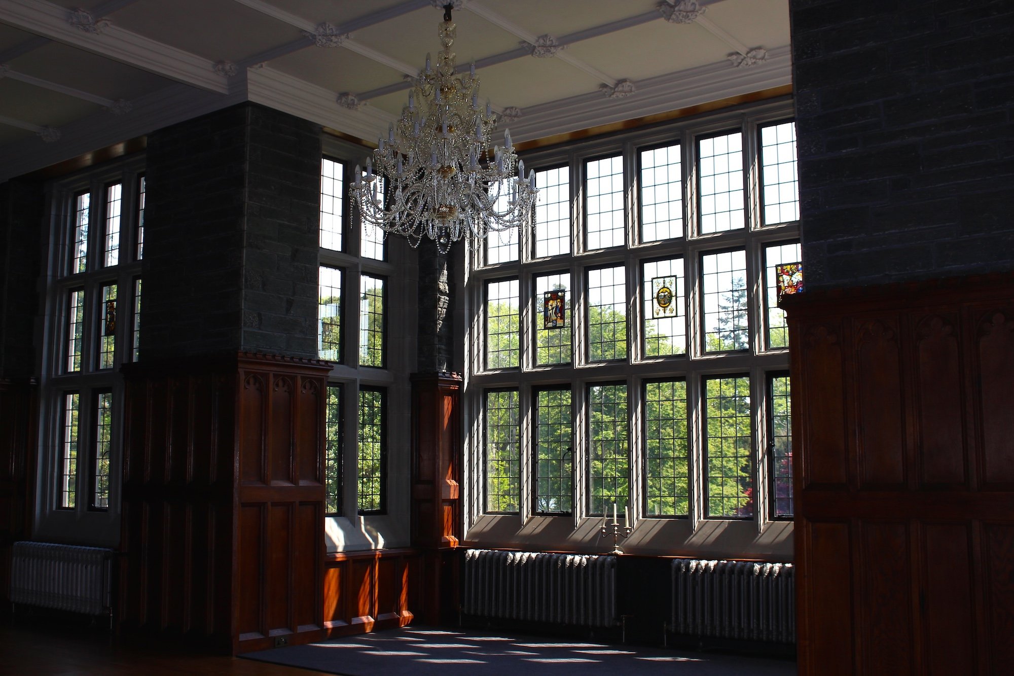 Interior of a large room with tall bay windows, stained glass accents, wooden paneling, and a grand chandelier hanging from a decorative ceiling.