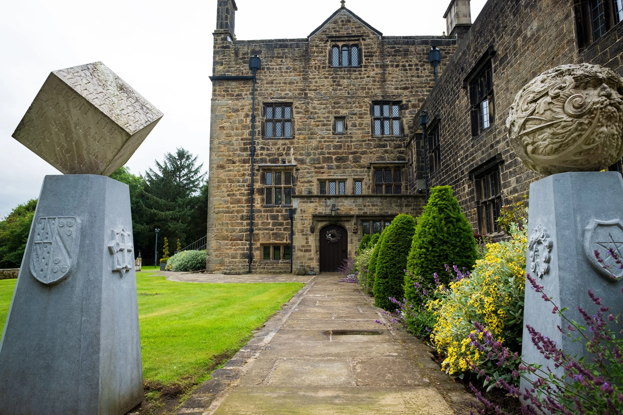 Stone building with gothic windows and a garden path, decorated with carved stone lion head wall ornament and stone sculptures with shields, surrounded by flowers and greenery.