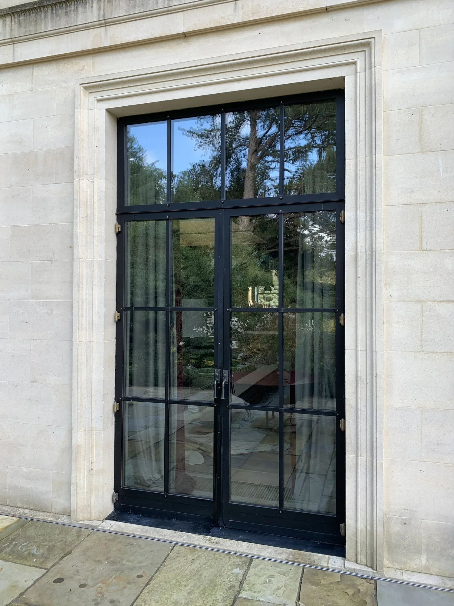 A black metal-framed glass door set in a beige stone wall, with a reflection of trees and the sky visible in the glass.