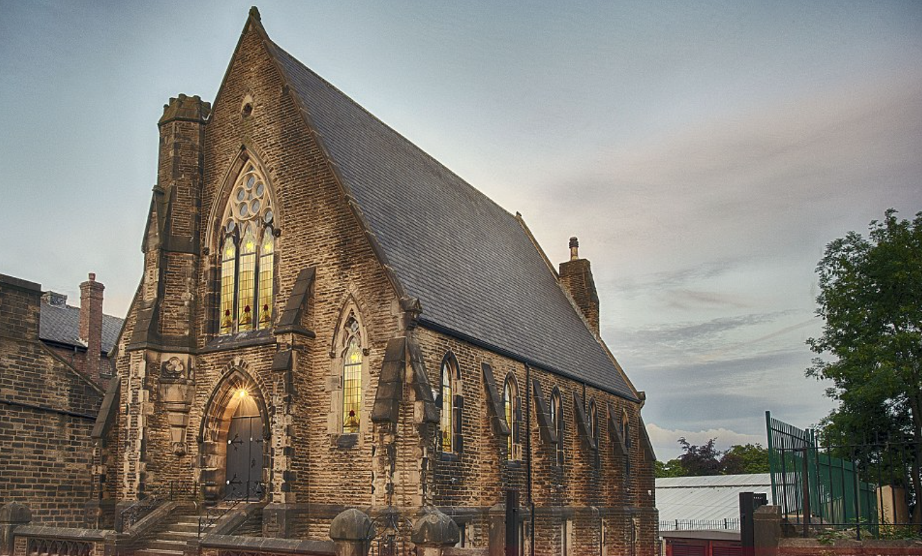 A Gothic-style brick church with stained glass windows and a steep roof, surrounded by a fence and trees, under a cloudy sky.