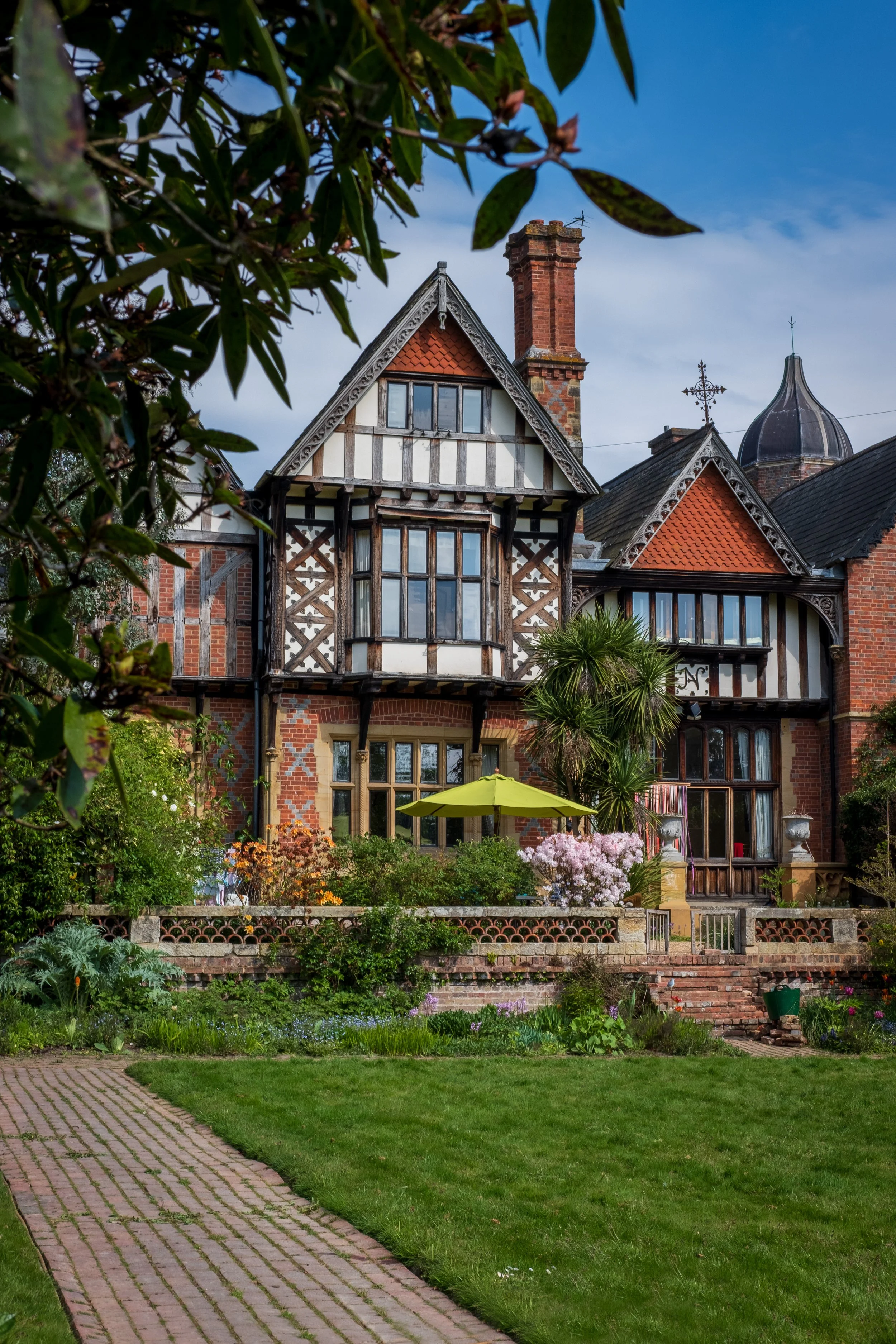 A large, historic, Tudor-style house with a garden in the foreground. The house features a steeply pitched roof, half-timbered walls, brick chimney, and decorative elements. The garden has blooming flowers, lush greenery, a yellow umbrella, and a pav