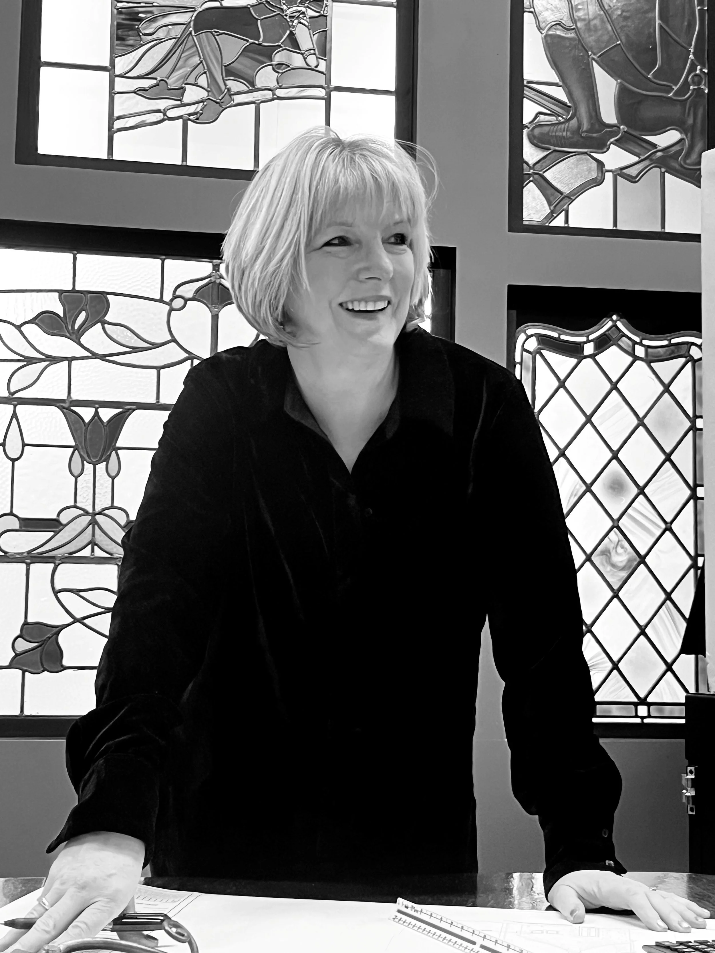 A smiling woman with short blond hair leaning on a table with paperwork and drawing tools, in front of decorative stained glass windows.
