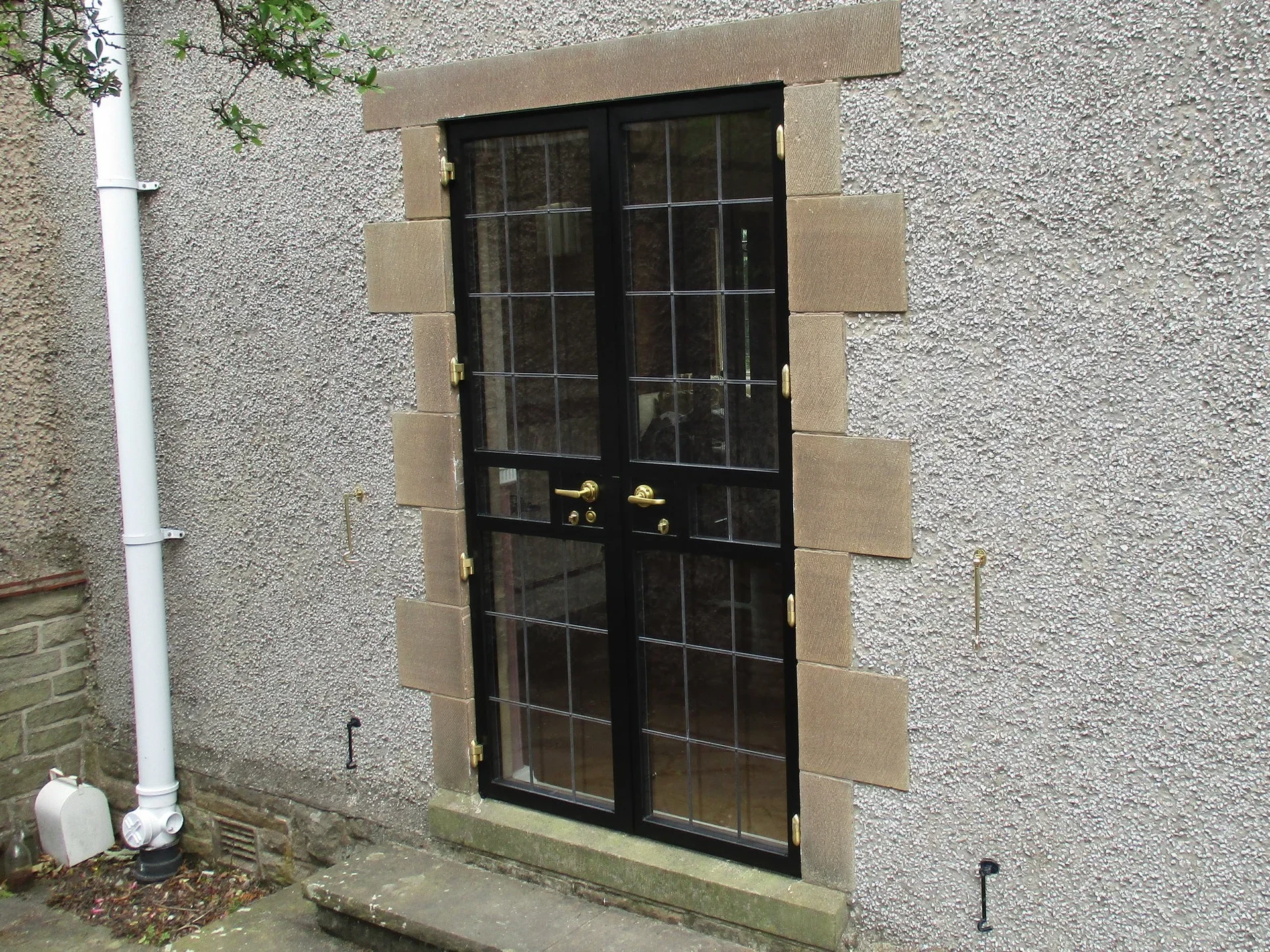 Exterior of a black metal door with glass panes, set in a beige stone frame on a textured gray wall. There are brass hinges and mail slot on the door, with a stone step at the base.