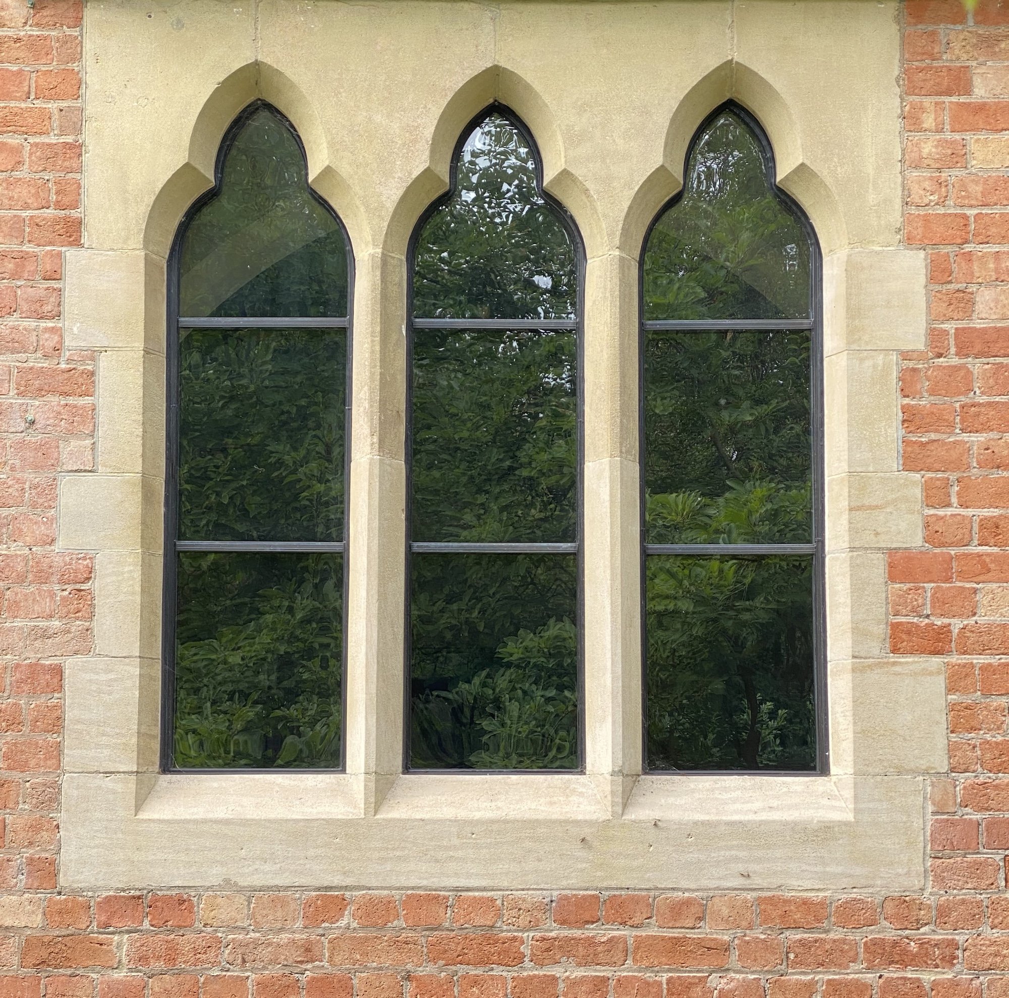 A wall with three Gothic-style arched windows, surrounded by a frame of cream-colored stone, embedded in a section of beige stucco on either side with red brick walls.