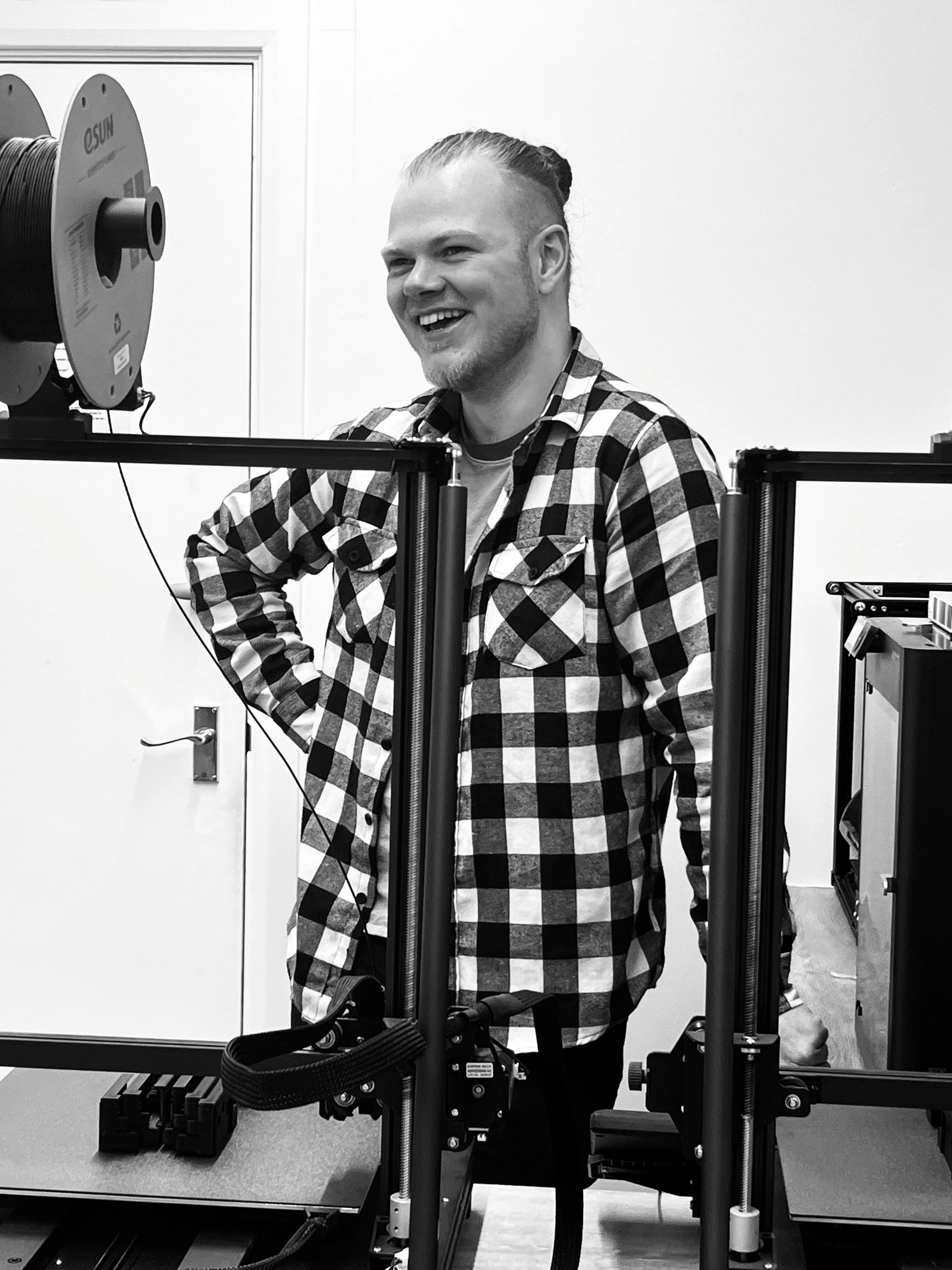 A man with a beard and long hair tied back, wearing a checkered shirt, standing and smiling in a room with equipment, possibly a 3D printer or CNC machine.