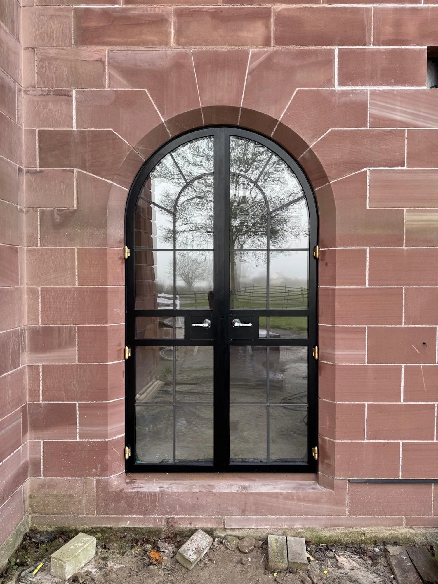 A black arched metal door with glass panes, set in a reddish-brown brick wall. Reflects trees and a foggy landscape outdoors.
