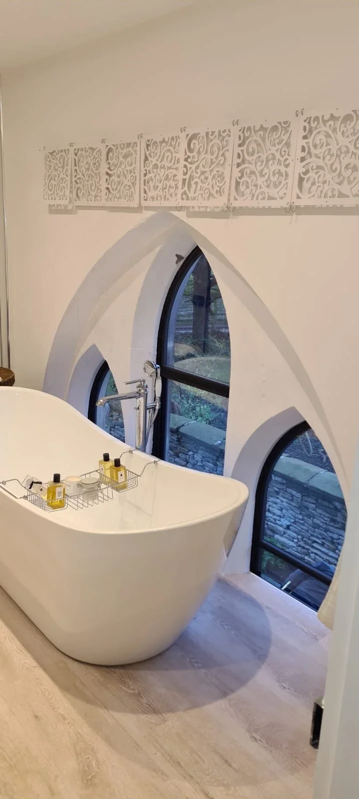 A modern bathroom with a freestanding bathtub under uniquely shaped arched windows, and decorative white lattice panels on the wall.