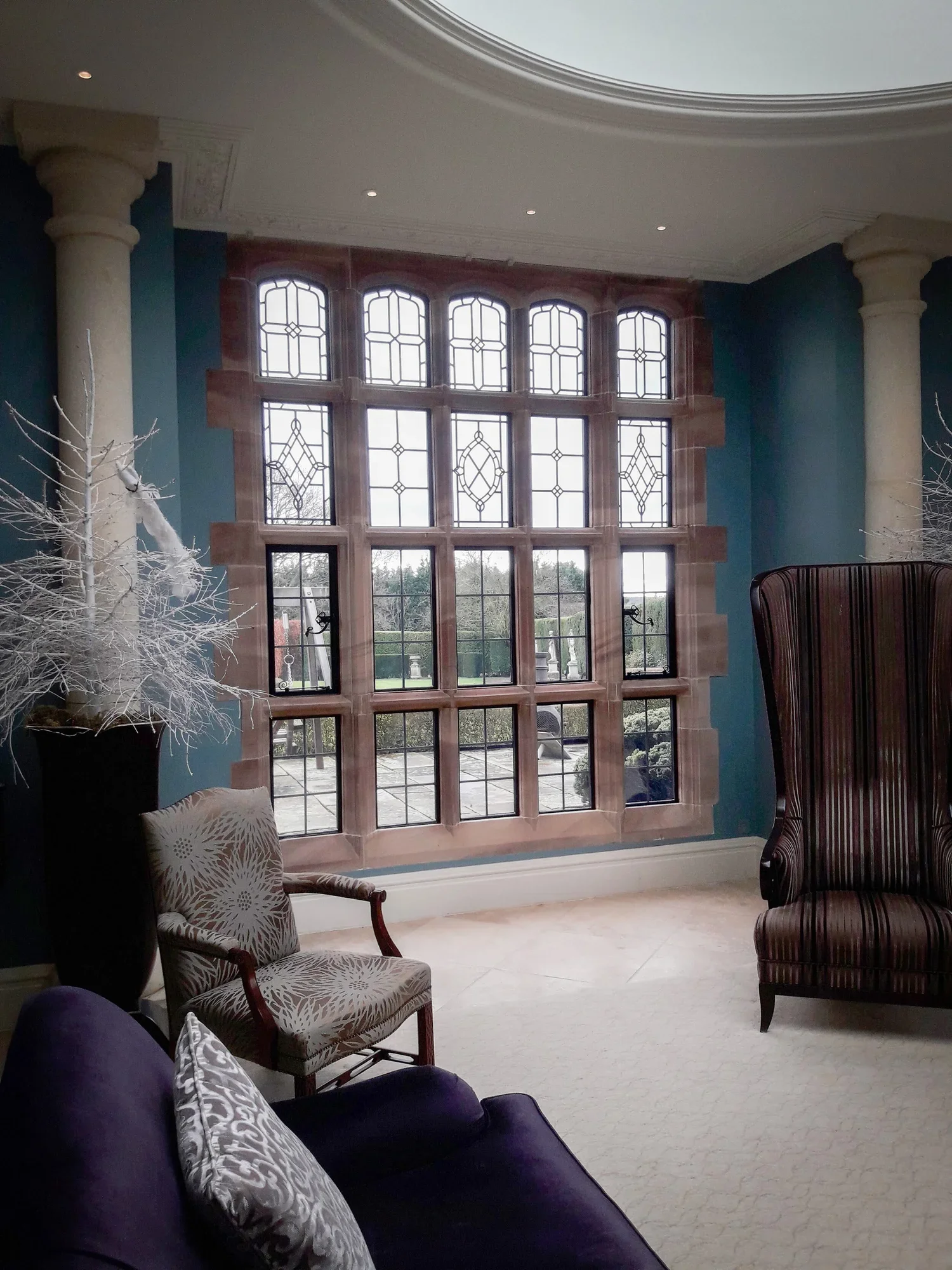 Living room with large decorative window, cream carpeted floor, purple sofa with patterned cushions, armchair near window, and decorative columns.