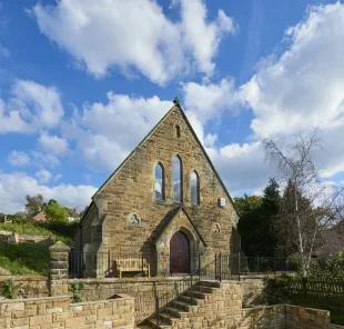 Stone church with pointed arch windows and door, under a partly cloudy sky, surrounded by trees and stone landscaping.