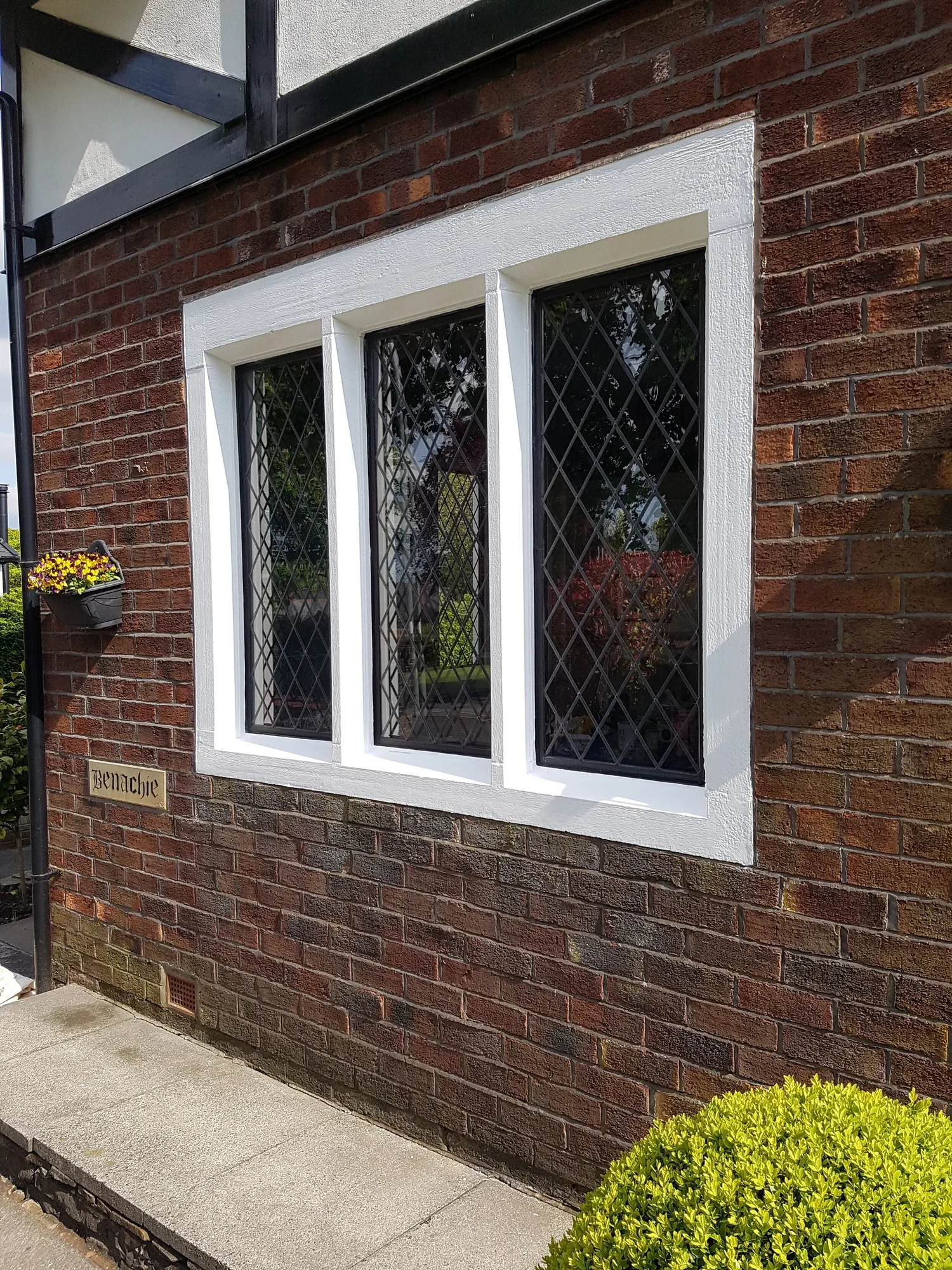Close-up of a brick house wall with three black-framed windows secured with black lattice bars. A small planter with yellow and purple flowers hangs on the wall. There is a nameplate reading "Benachie" below the window along with a small drainage ven
