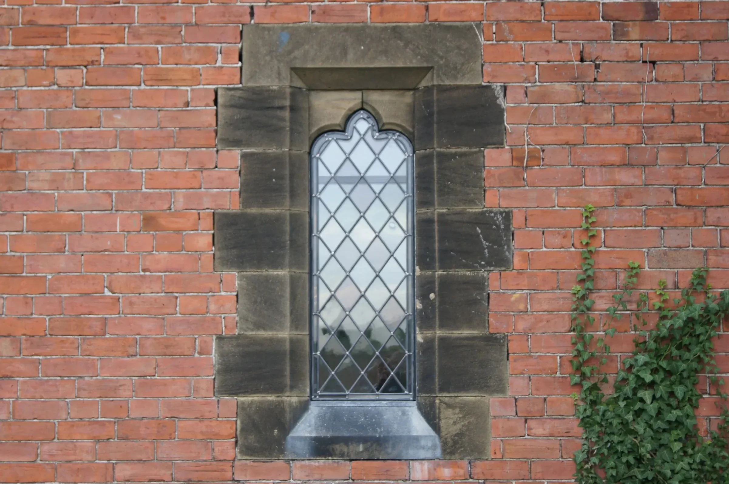A small, narrow window with a pointed, arch-shaped top, set into a brick wall with a stone frame and a diamond-patterned metal grille, and some green plants growing on the right side.