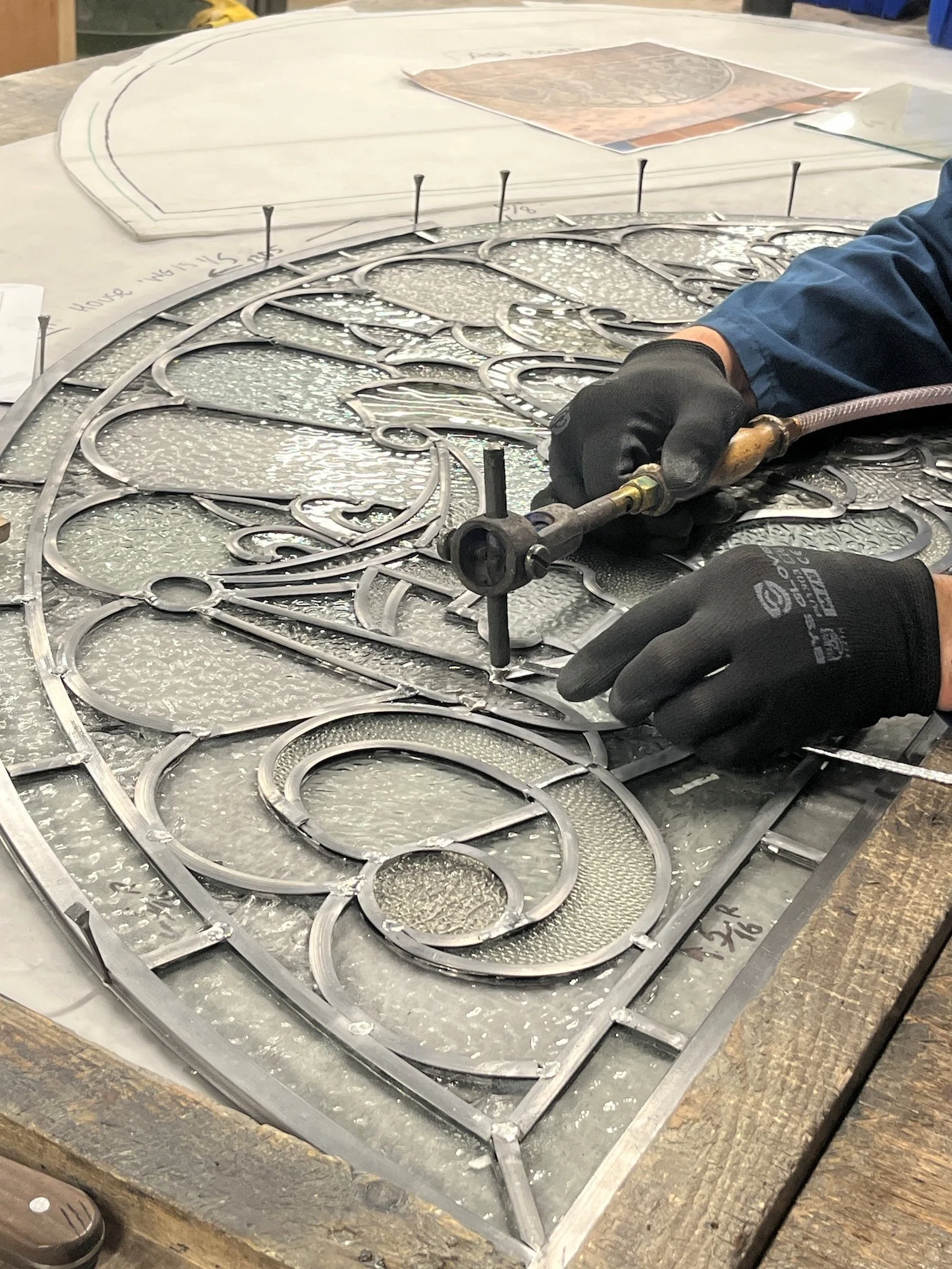 Individual working on a decorative metal piece with intricate design, using a tool while wearing black gloves in a workshop.