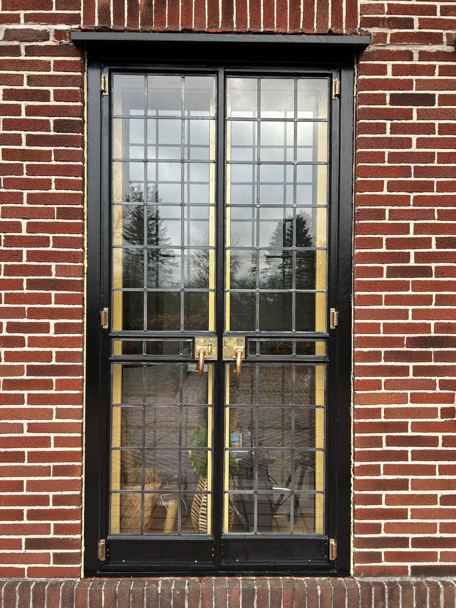 Black metal door with glass panels in a red brick wall, reflecting trees and cloudy sky.