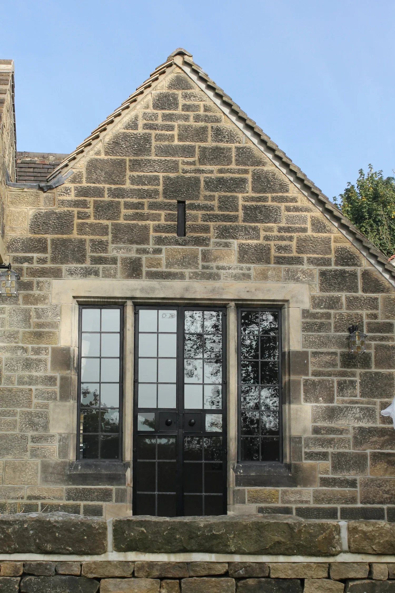 Front view of a stone house with a gabled roof, black framed glass doors, and tall narrow windows on either side, set on a stone foundation.