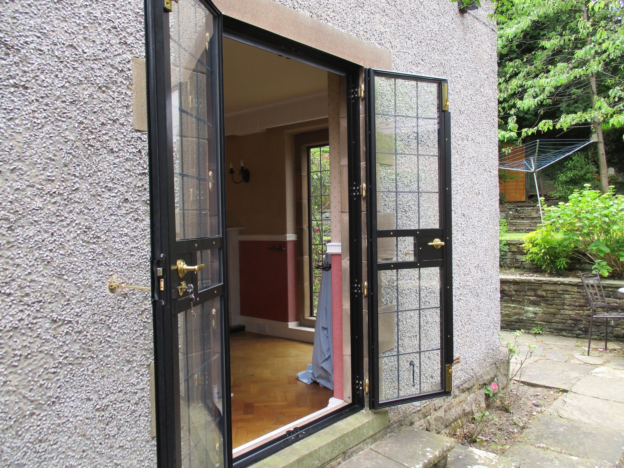 Open black metal window with glass panes on a textured exterior wall, revealing a room with wooden floors and a beige wall inside. A cloth is hanging outside the window, and green trees and plants are visible in the background.