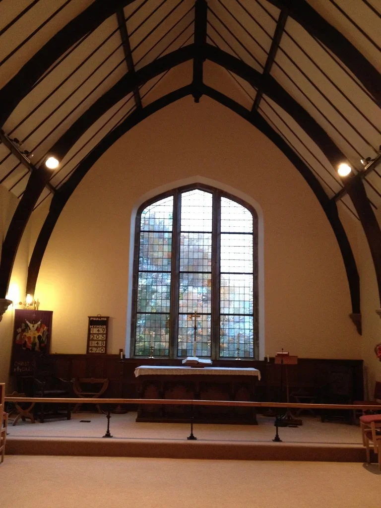 Interior of a church with an altar, a large stained glass window, and wooden beams on the ceiling.