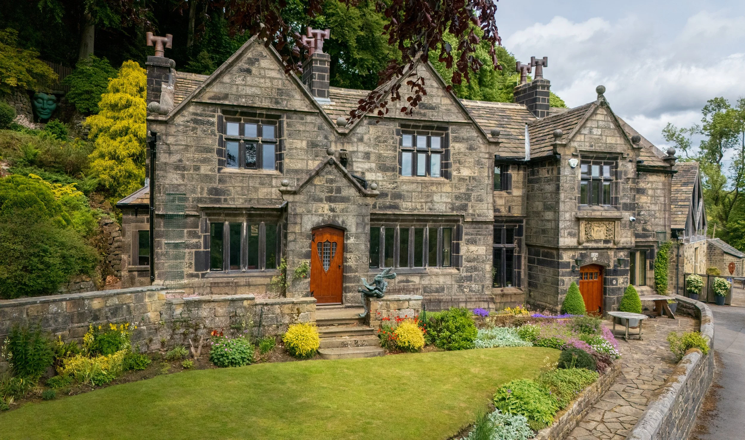 Stone house with multiple chimneys and large windows, surrounded by colorful garden with flowers, trees, and shrubs, under a cloudy sky.