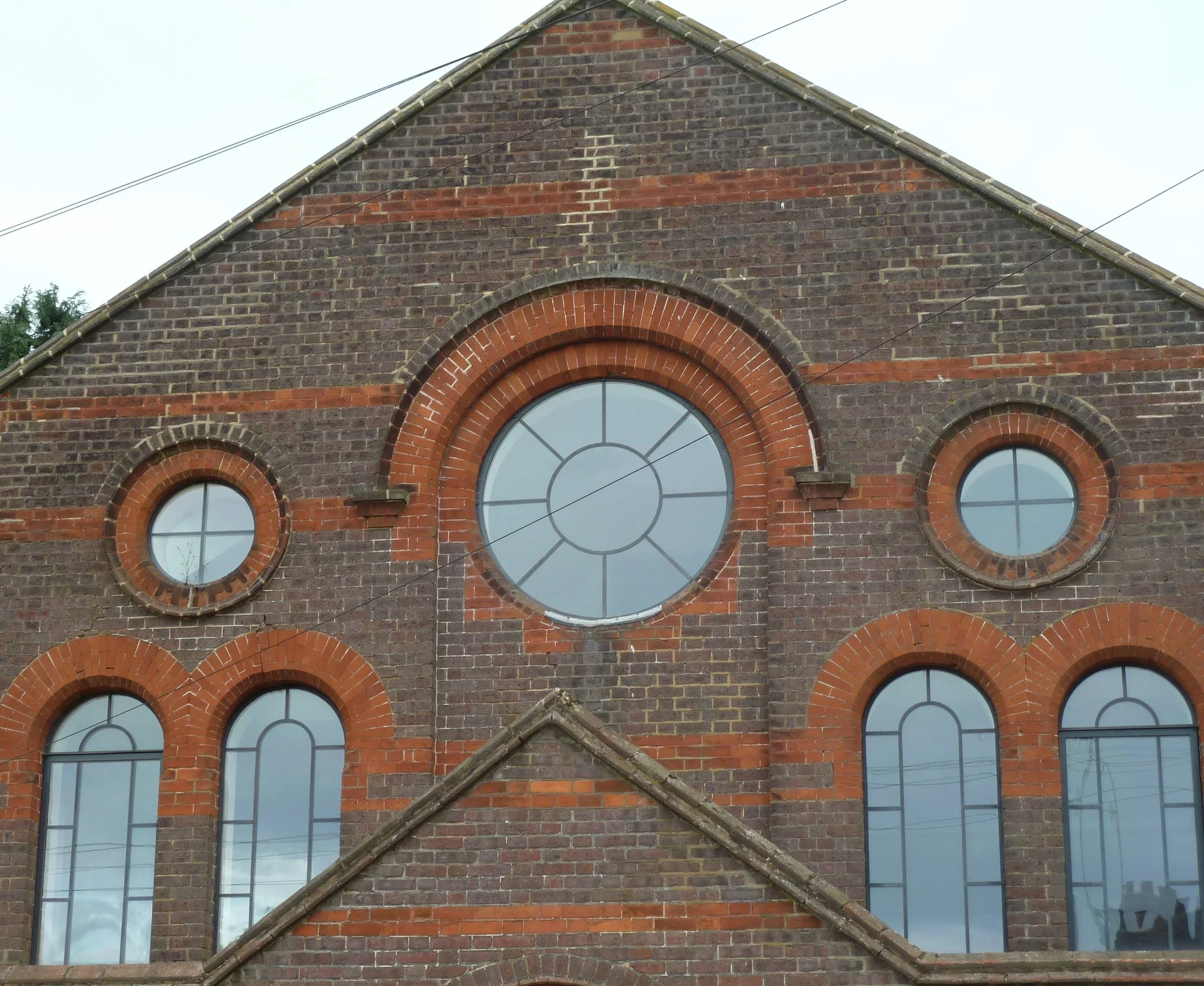 Close-up of a red brick building with large arched windows, some of which have a circular design in the center, and power lines running across the top.