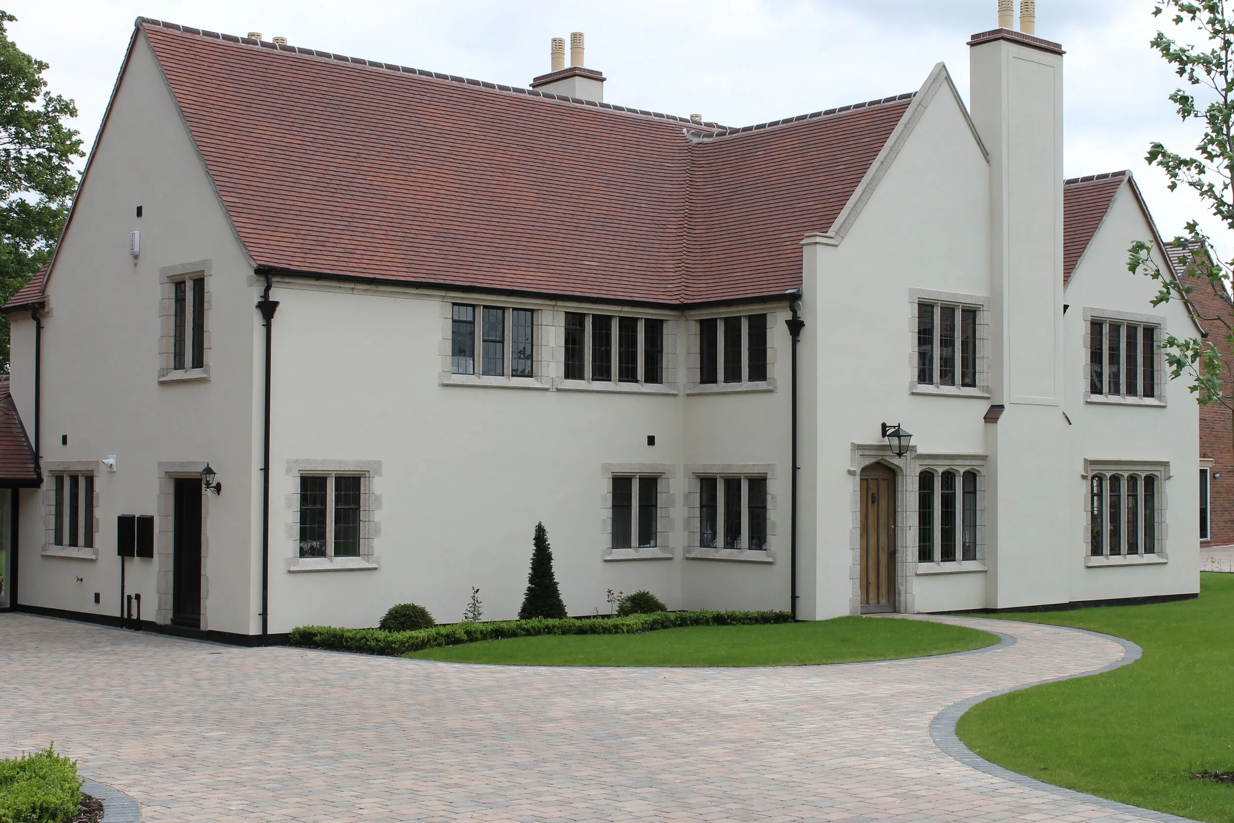 A large white house with a red-tiled roof, multiple chimneys, and several windows, surrounded by a neatly landscaped yard with a paved walkway and grass.