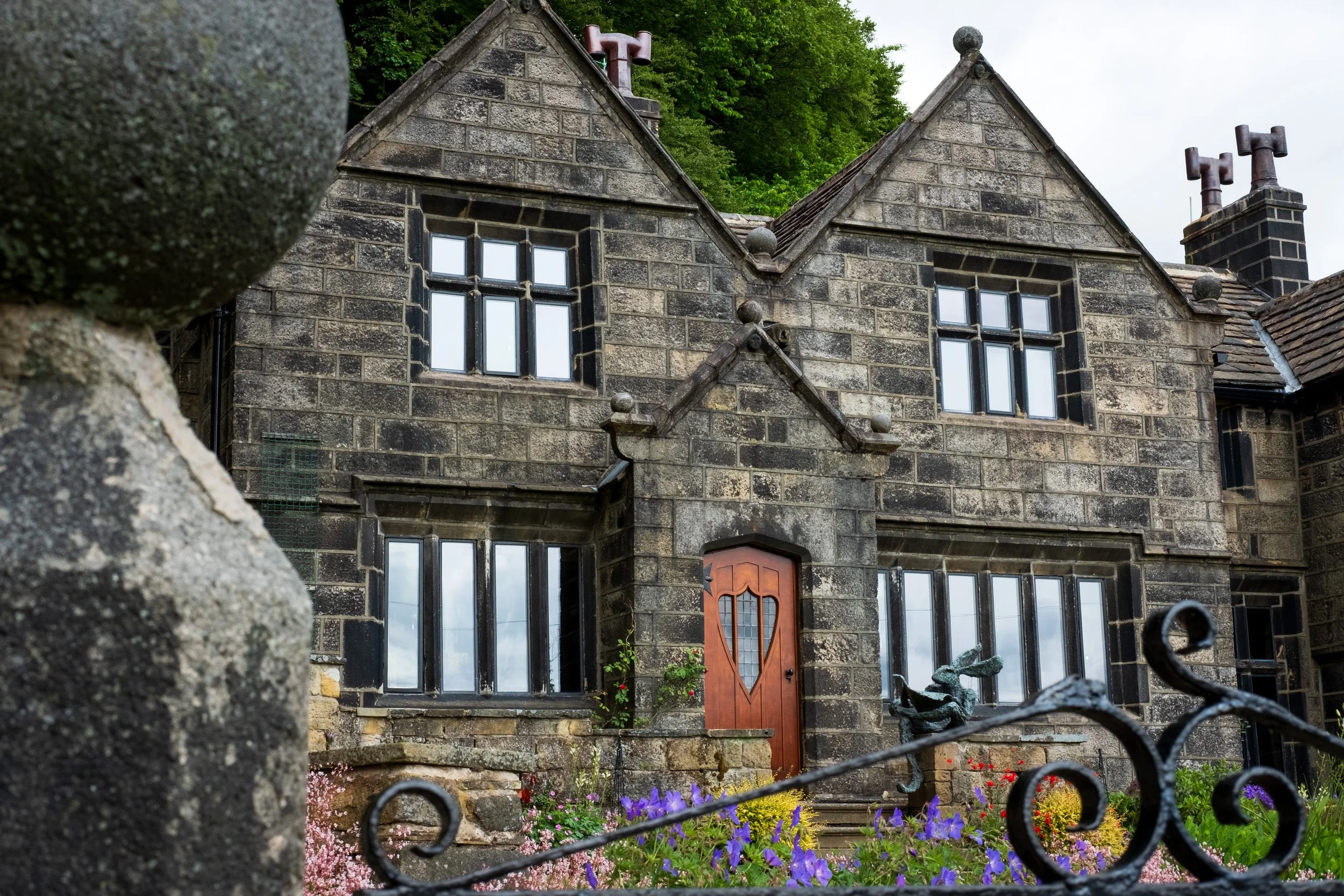 View of a historic stone house with multiple gabled roofs, small windows, and a wooden front door, surrounded by colorful flowers and partially obscured by a stone wall and wrought iron fence in the foreground, with a background of green trees and an