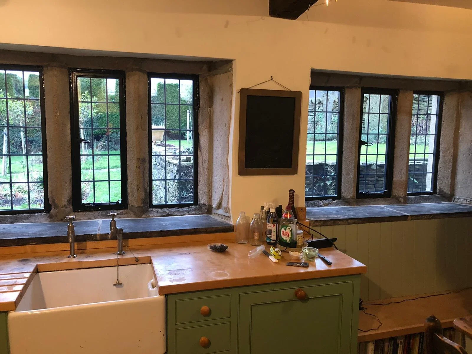 A rustic kitchen with four large windows looking out onto a garden. The kitchen features a farmhouse sink with a wooden countertop, some bottles, and cleaning supplies on the counter. There's a blackboard hanging on the wall between the windows.