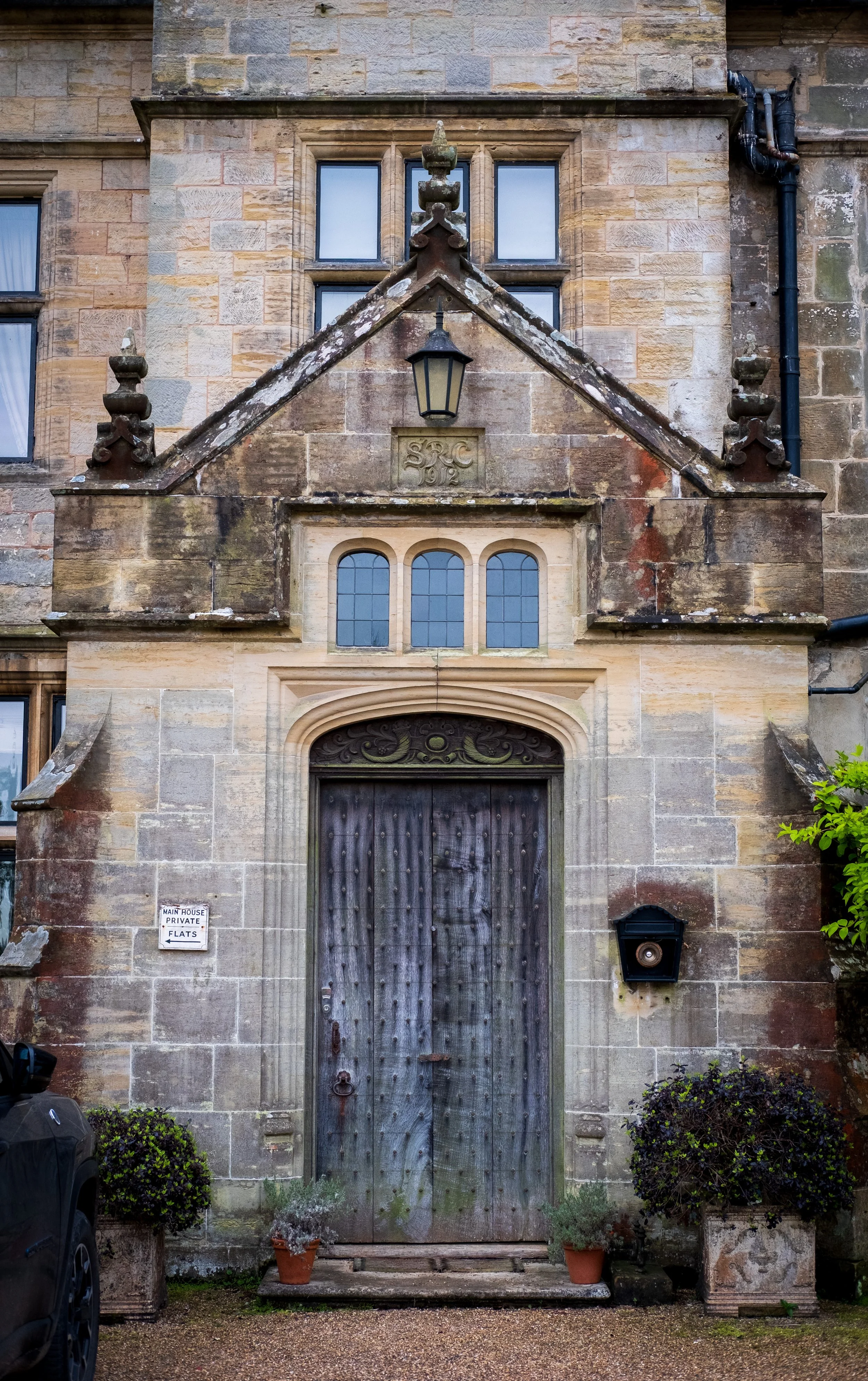 An old stone building with a wooden door, decorative stonework, a lamp hanging above the door, a sign indicating 'Main House Private Flats,' and potted plants on either side of the entrance.