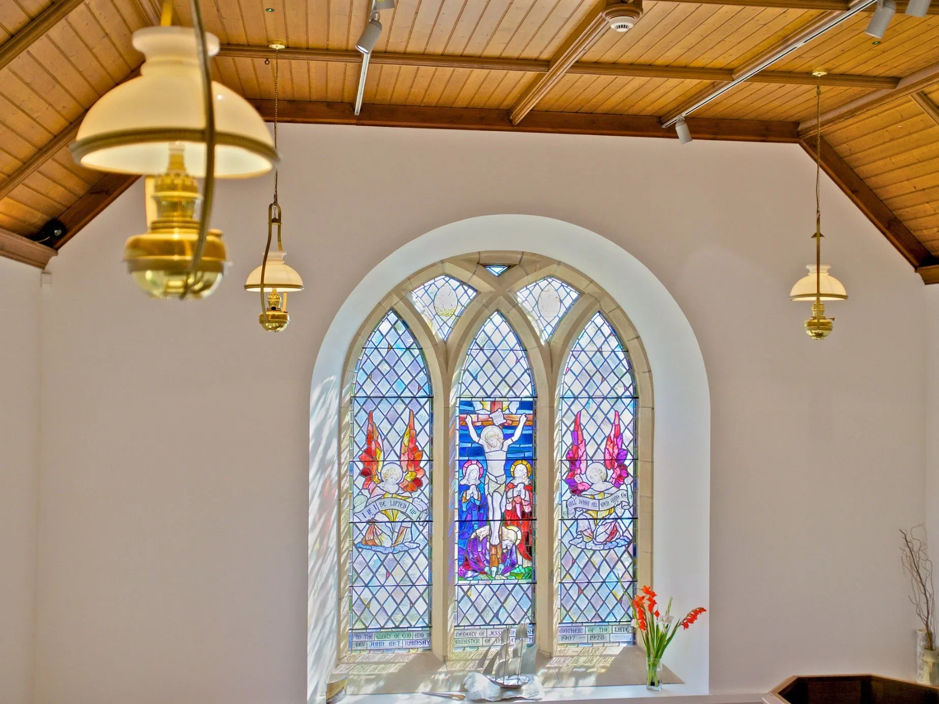 Sunlit stained glass window featuring religious figures and inscriptions, located inside a church with wooden ceiling, hanging lamps, and a small potted plant with orange flowers below.