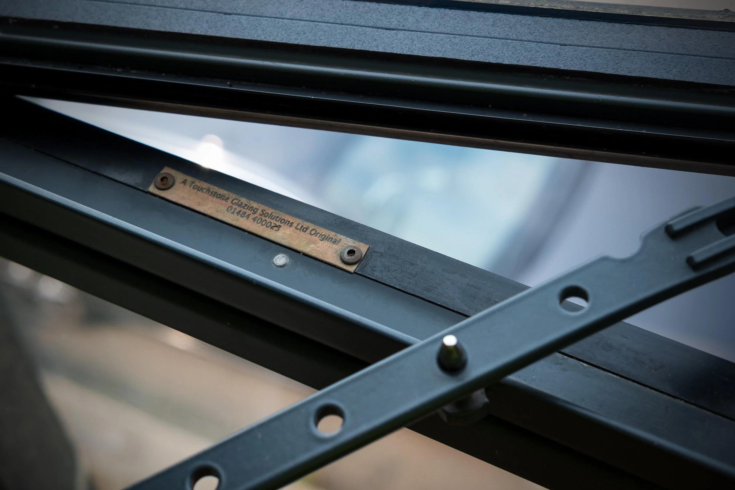Close-up of a metal frame with a label reading, 'A Touchstone Glazing Solutions Ltd Original 01484 400023', attached to a partially assembled structure, with a cloudy sky visible through an opening.