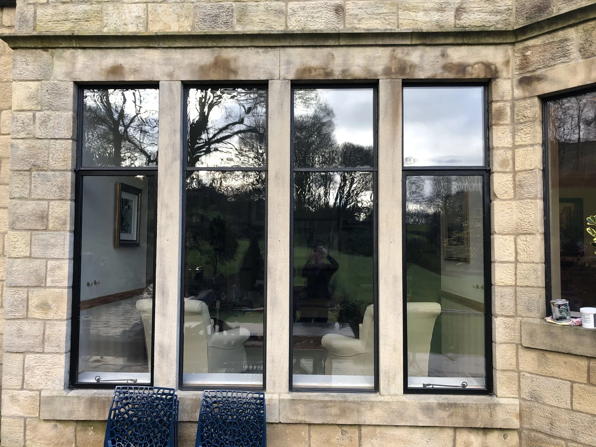 The image shows a stone building exterior with three large glass windows reflecting trees and the sky. Inside the room, visible through the windows, are a cream-colored armchair and some art on the walls. Outside the building, there are blue chairs a