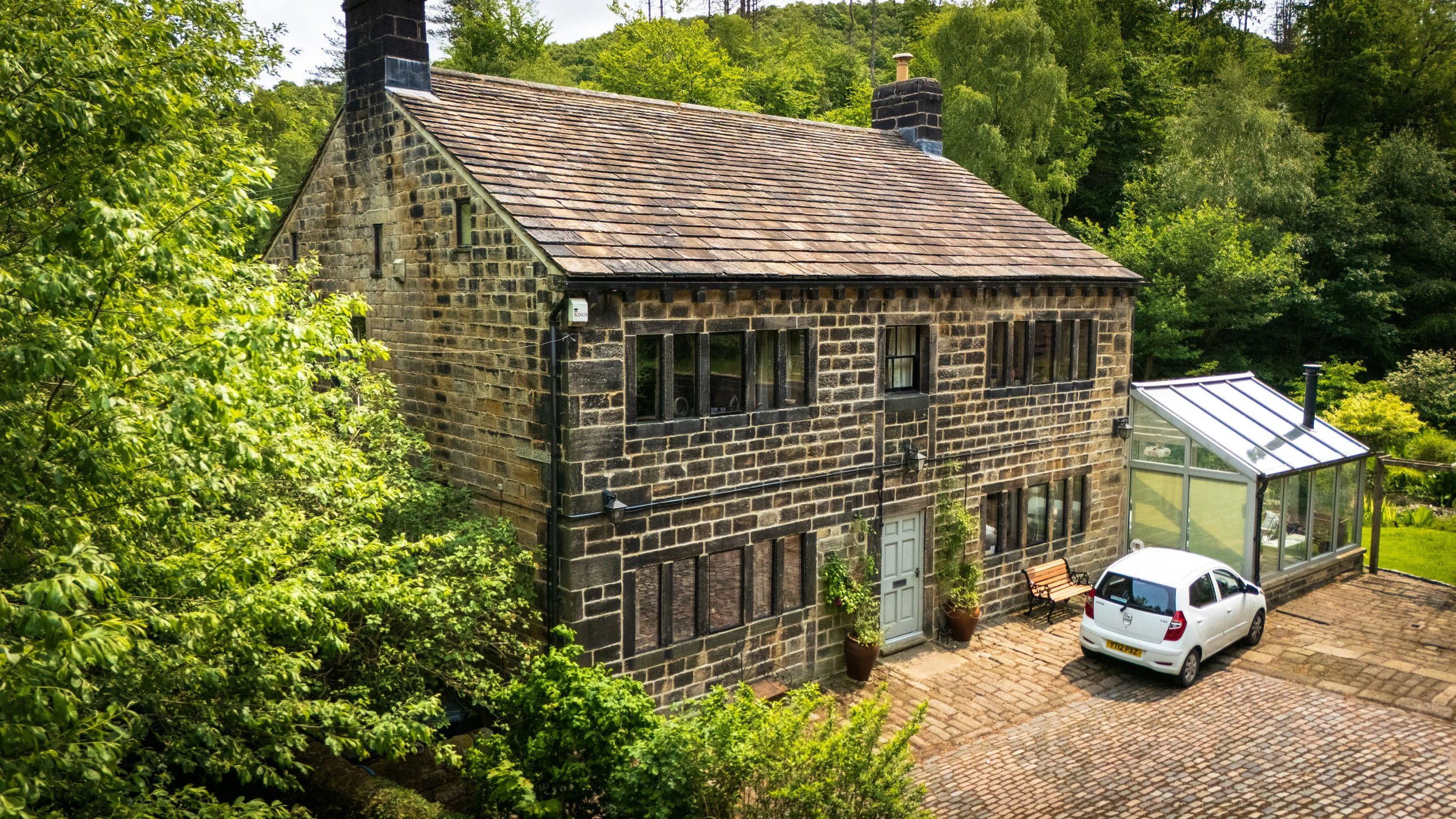 A stone house with a pitched roof, multiple windows, and a small glass greenhouse attached, surrounded by green trees and a paved courtyard with a bench and a white car.