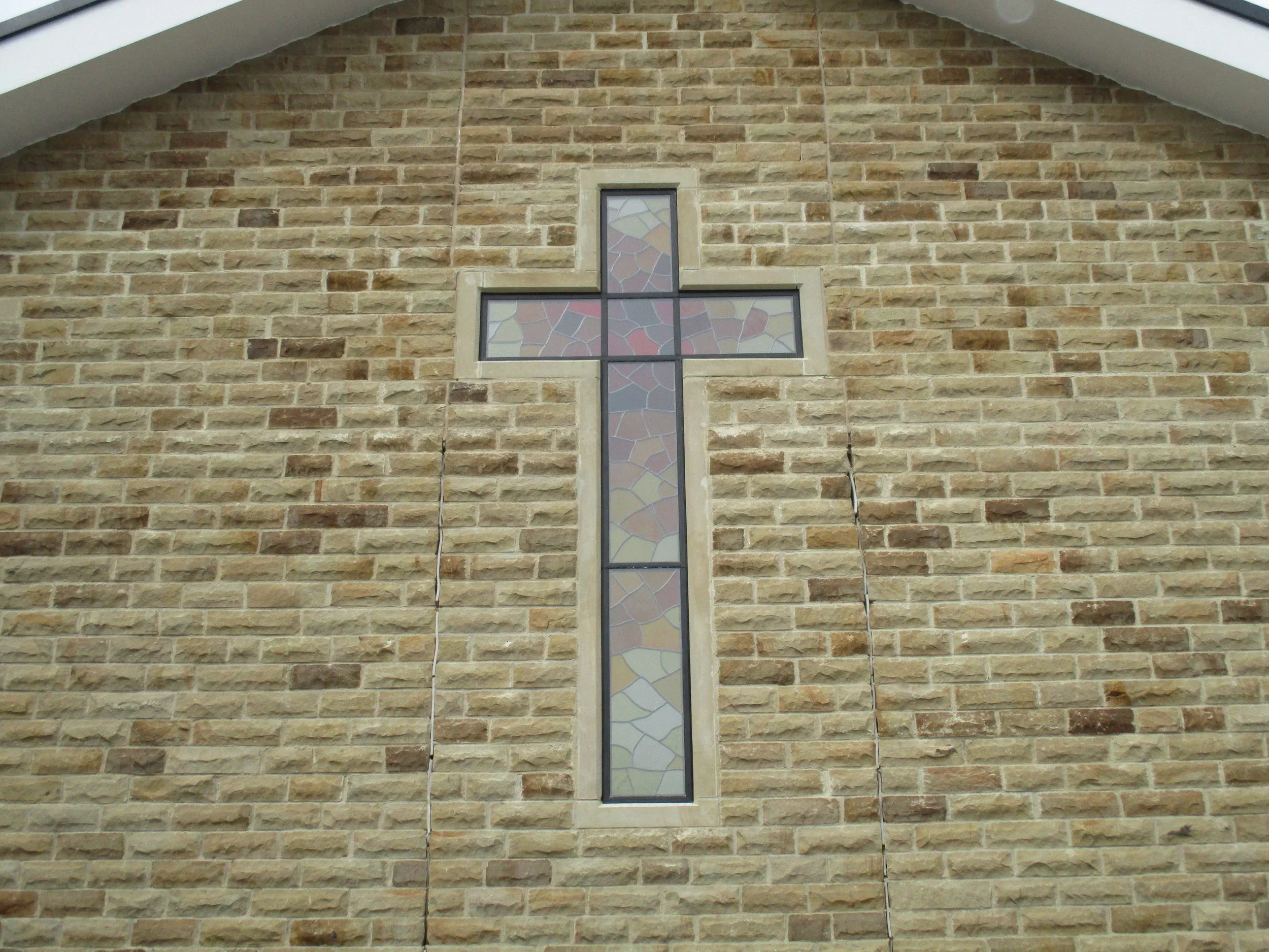 Interior wall of a building made of light brown bricks with a stained glass window in the shape of a cross.