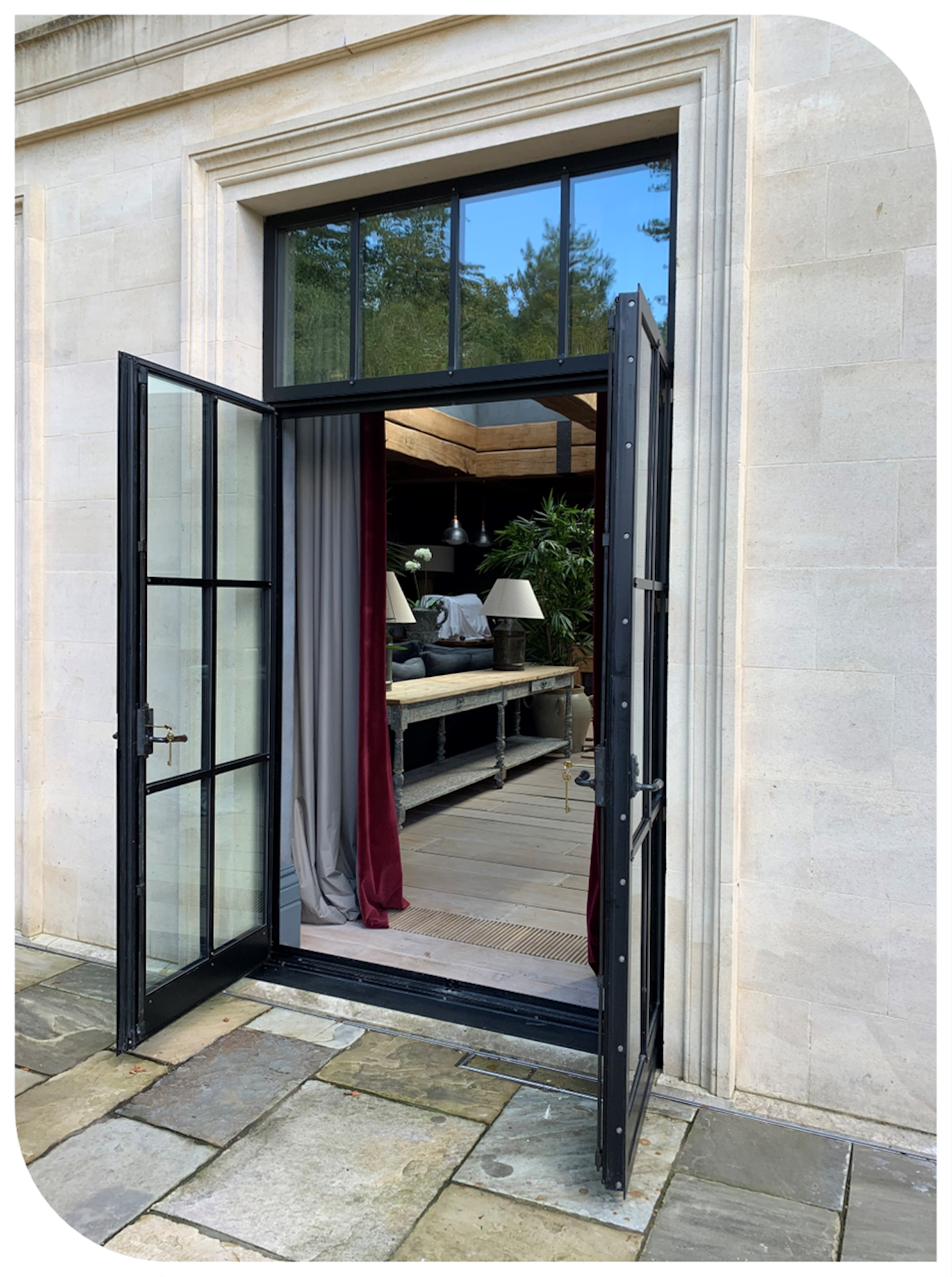 Open black-framed glass door leading from stone patio into a living room with wooden ceiling beams, hanging pendant lights, and houseplants.
