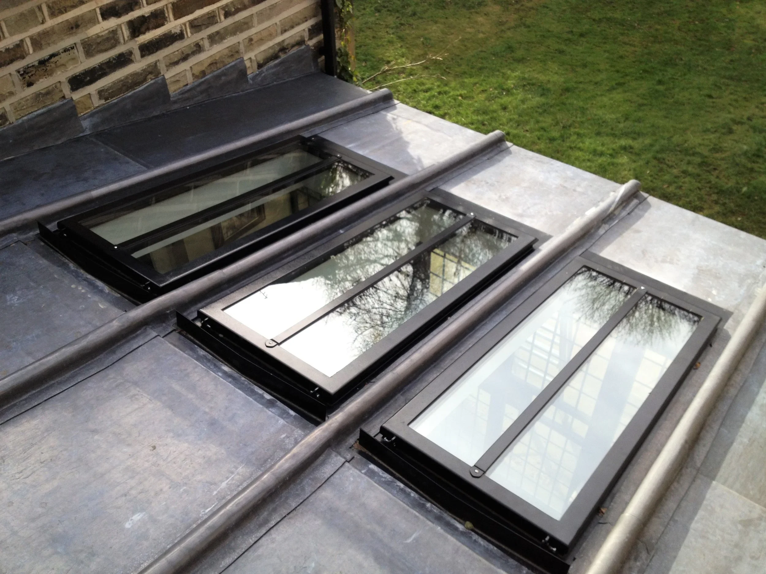 Three roof skylights with black frames and glass panes on a flat rooftop, with a grassy yard and trees reflected in the glass.