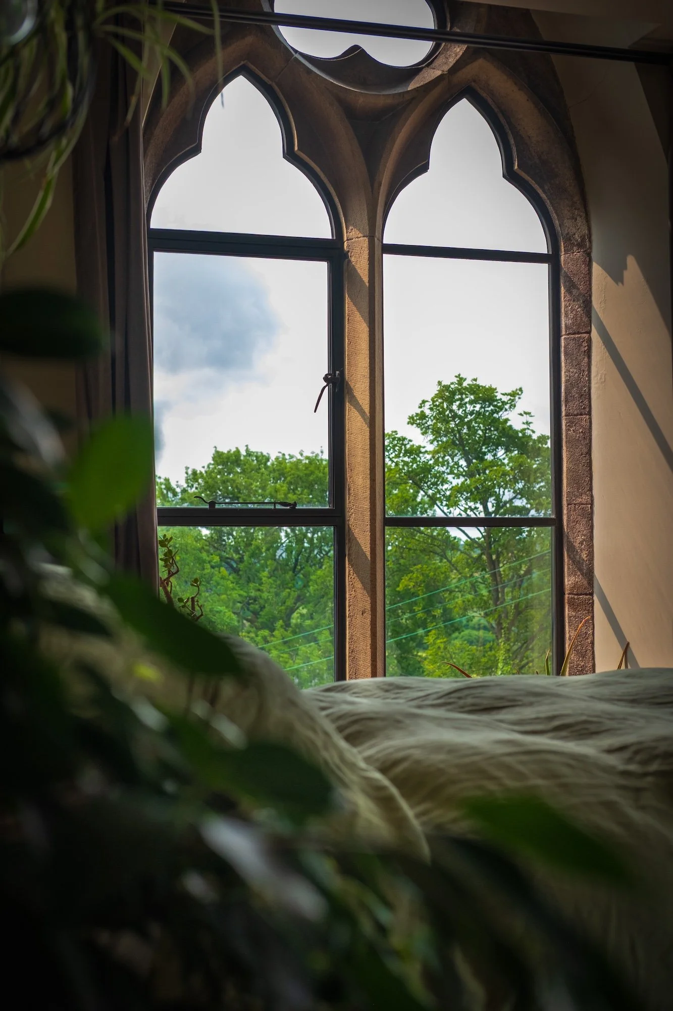 View from a bed looking out a window with gothic arch design, showing green trees outside on a cloudy day.