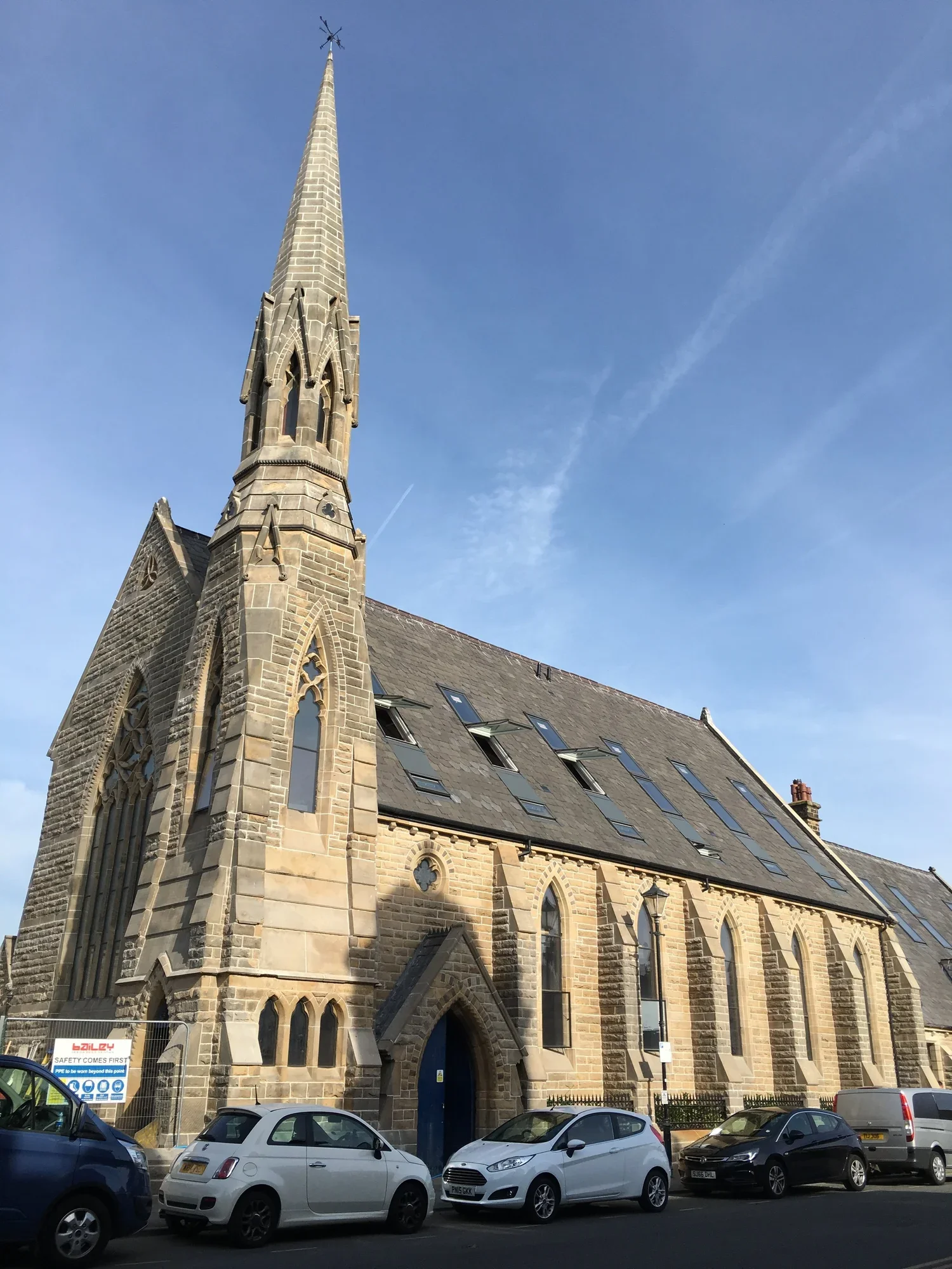 A historic stone church with a tall steeple topped with a weather vane, parked cars in the foreground, and a partly cloudy blue sky.
