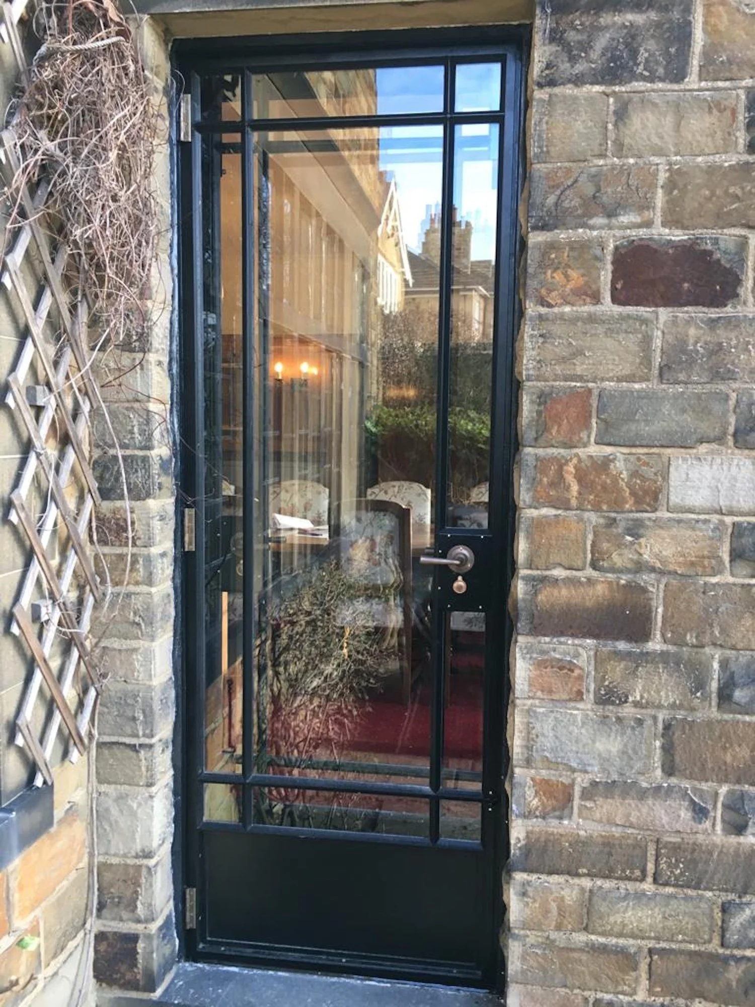 A black metal screen door with a handle, set into a historic brick wall with an inset stone frame, looking into a cozy interior with warm lighting, a red carpet, and some furniture visible inside.