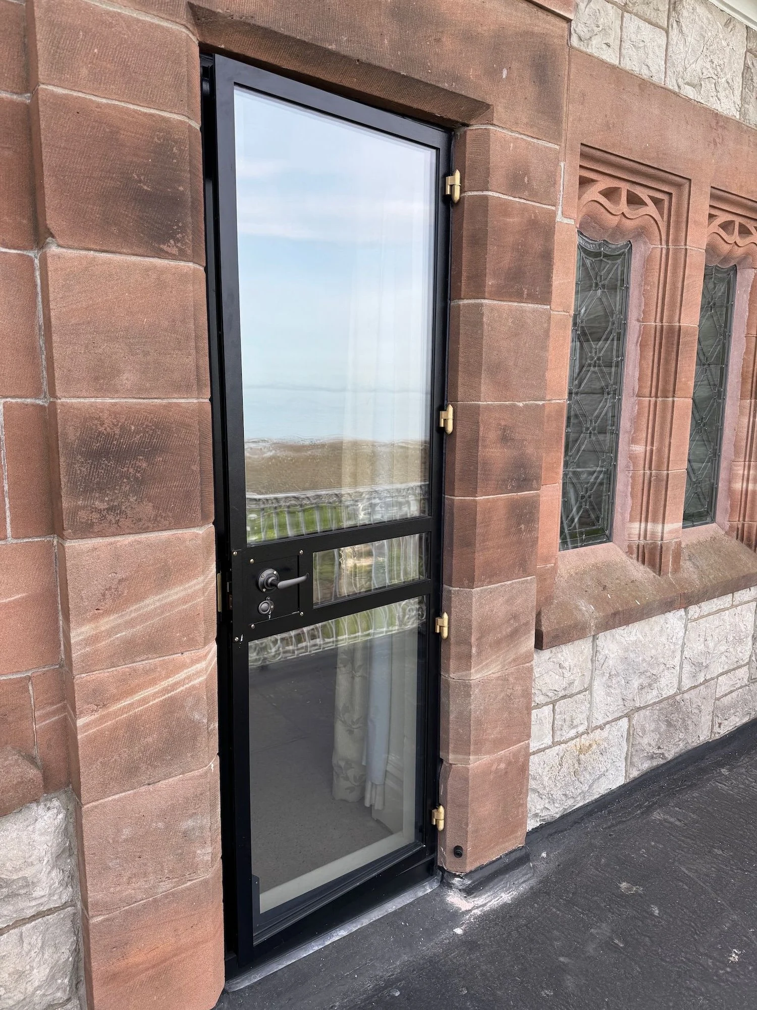 A black-framed glass door with brass hinges and a lock, set into a stone building with decorative stonework and stained glass windows, overlooking a landscape with green grass and a body of water.