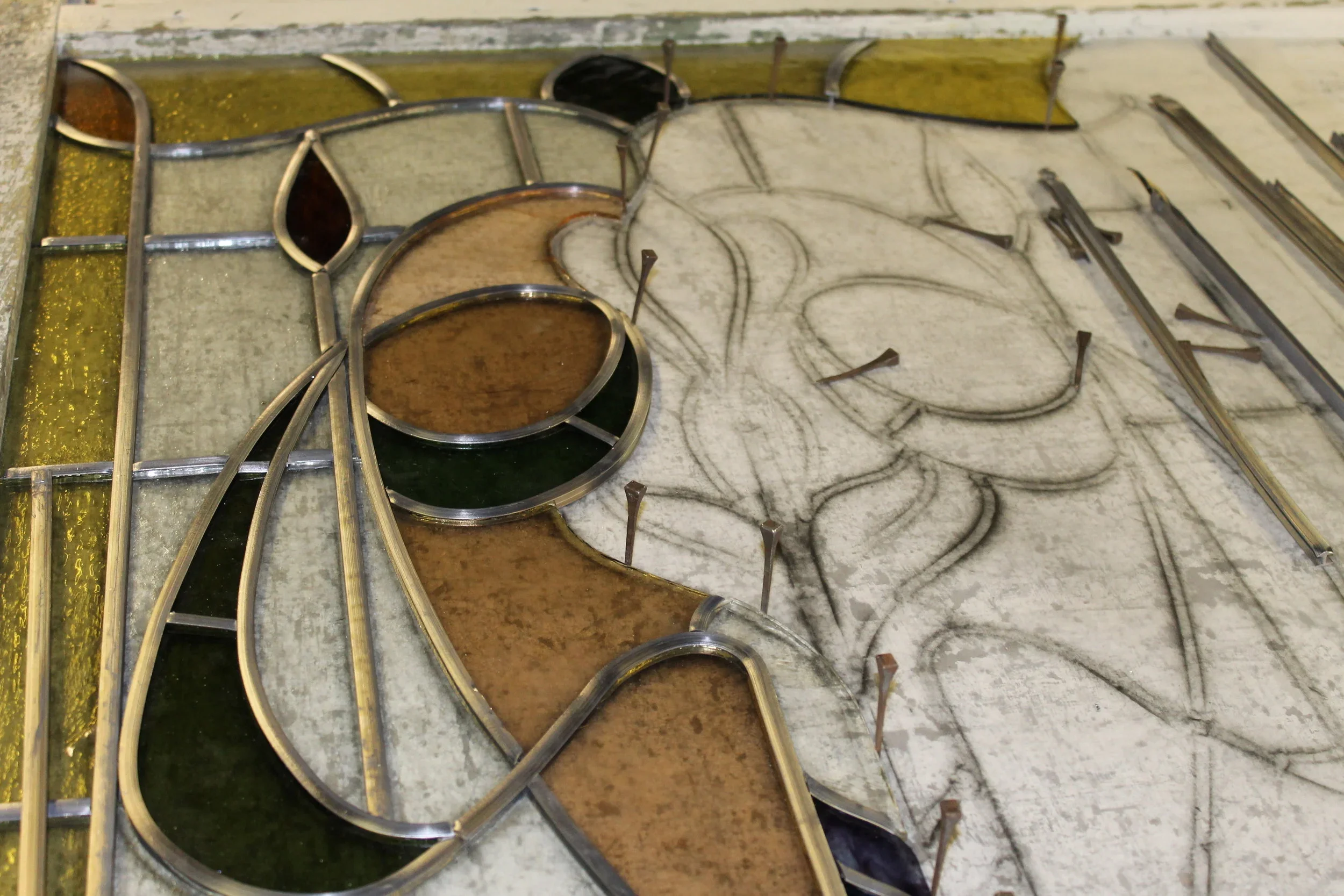 A stained glass window with a floral design in progress, featuring metal framework and some colored glass pieces in gold, black, and brown.